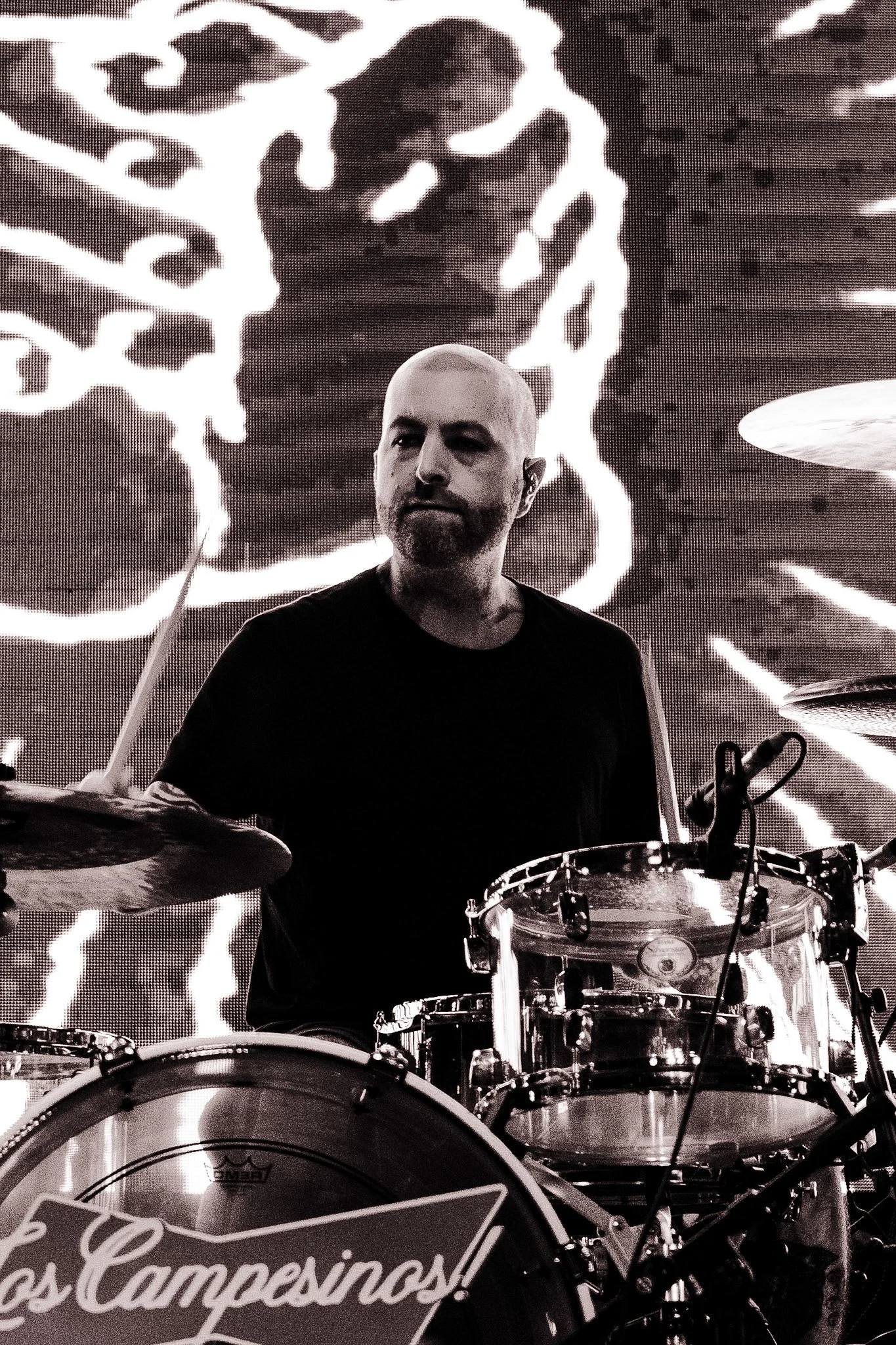 A man with a beard and shaved head playing drums on stage, with a large illuminated digital display in the background showing an image of a face and abstract patterns.