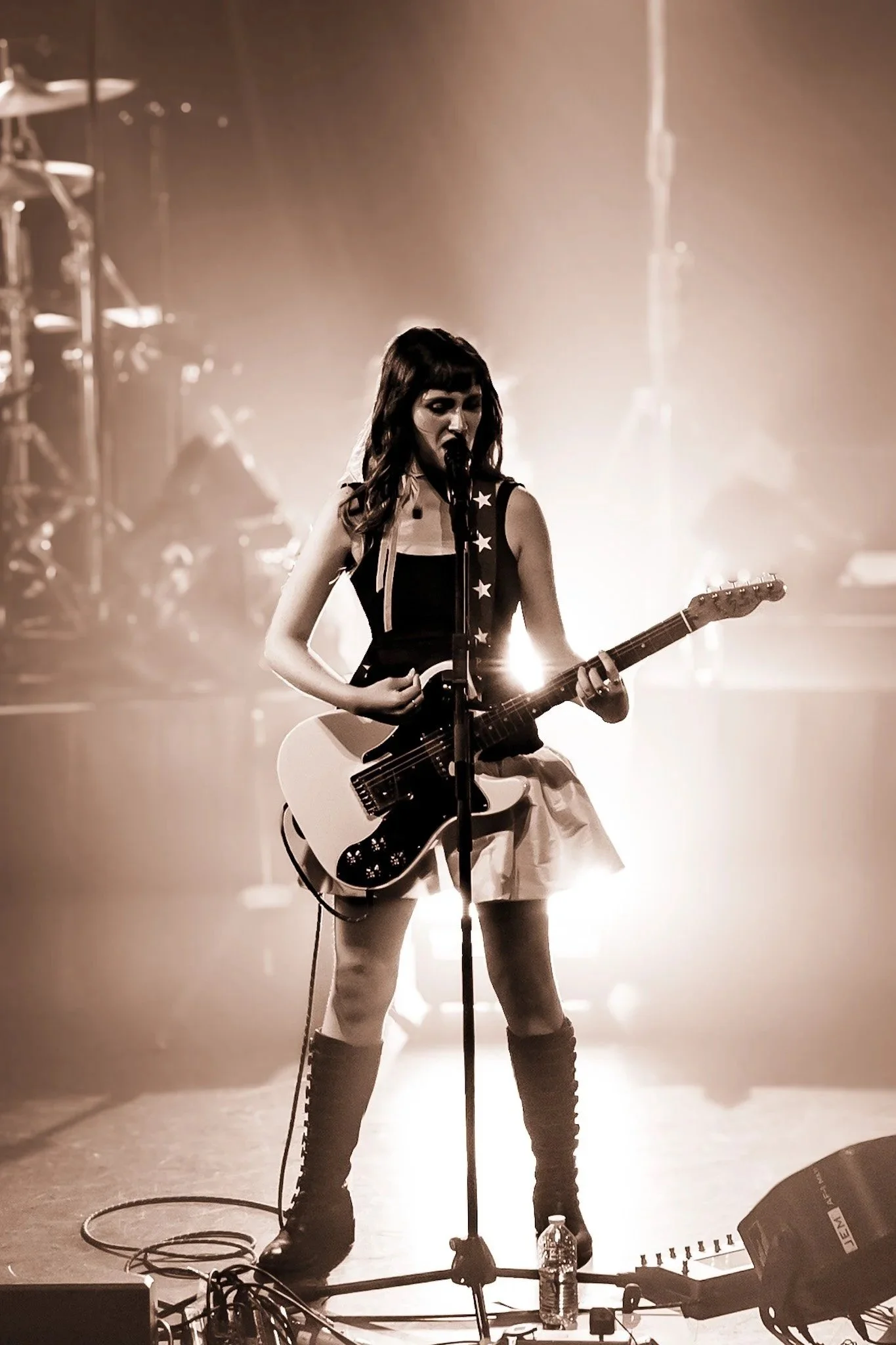 A female musician standing on stage, singing into a microphone and playing an electric guitar. She has shoulder-length hair and is wearing a sleeveless top, skirt, and knee-high boots. The stage lighting creates a bright, backlit effect.