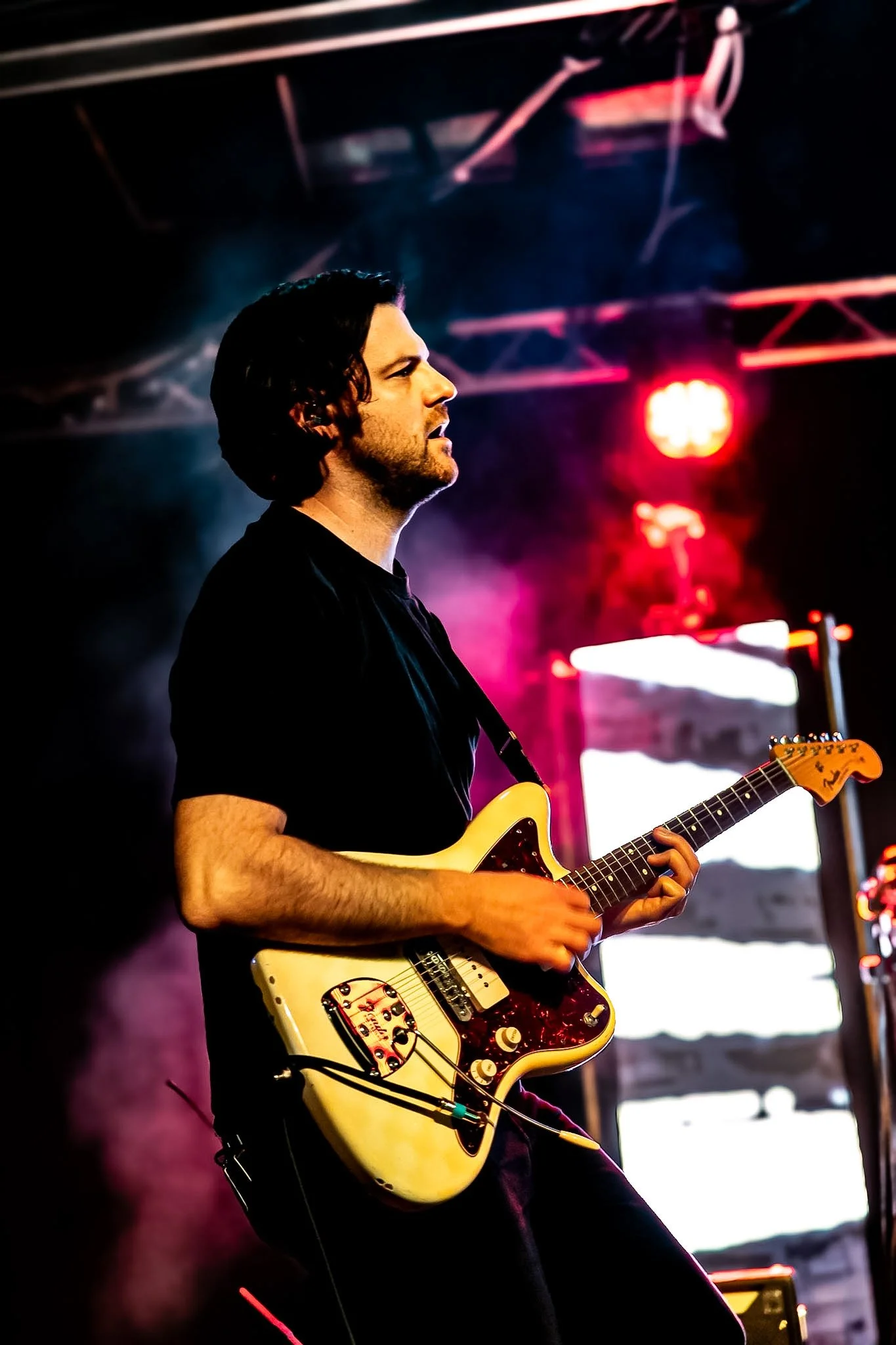 A male musician playing an electric guitar on stage, with colorful stage lighting and a dark background.