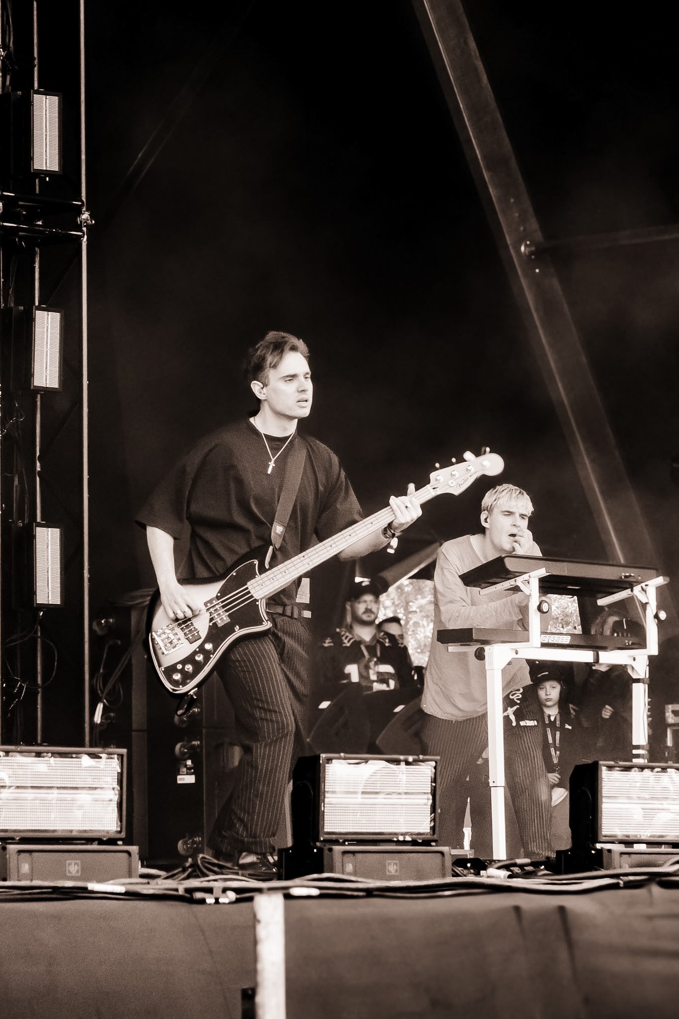 Black and white photo of young musicians performing on stage, with one person playing bass guitar and another singing or speaking into a microphone, surrounded by stage equipment and audience.
