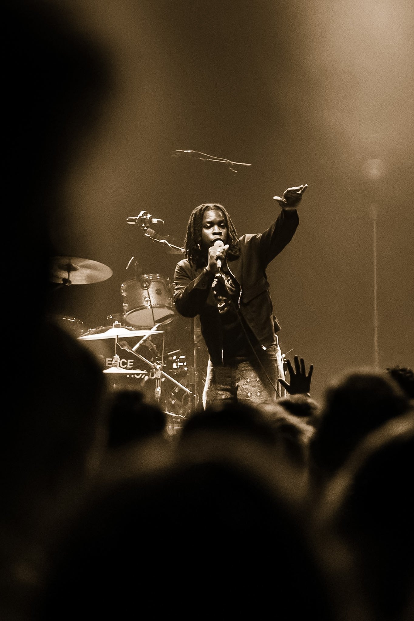 Black and white photo of a singer with dreadlocks on stage, holding a microphone and raising their hand, with a drum set and other musical equipment in the background, audience silhouettes and raised hands in the foreground.