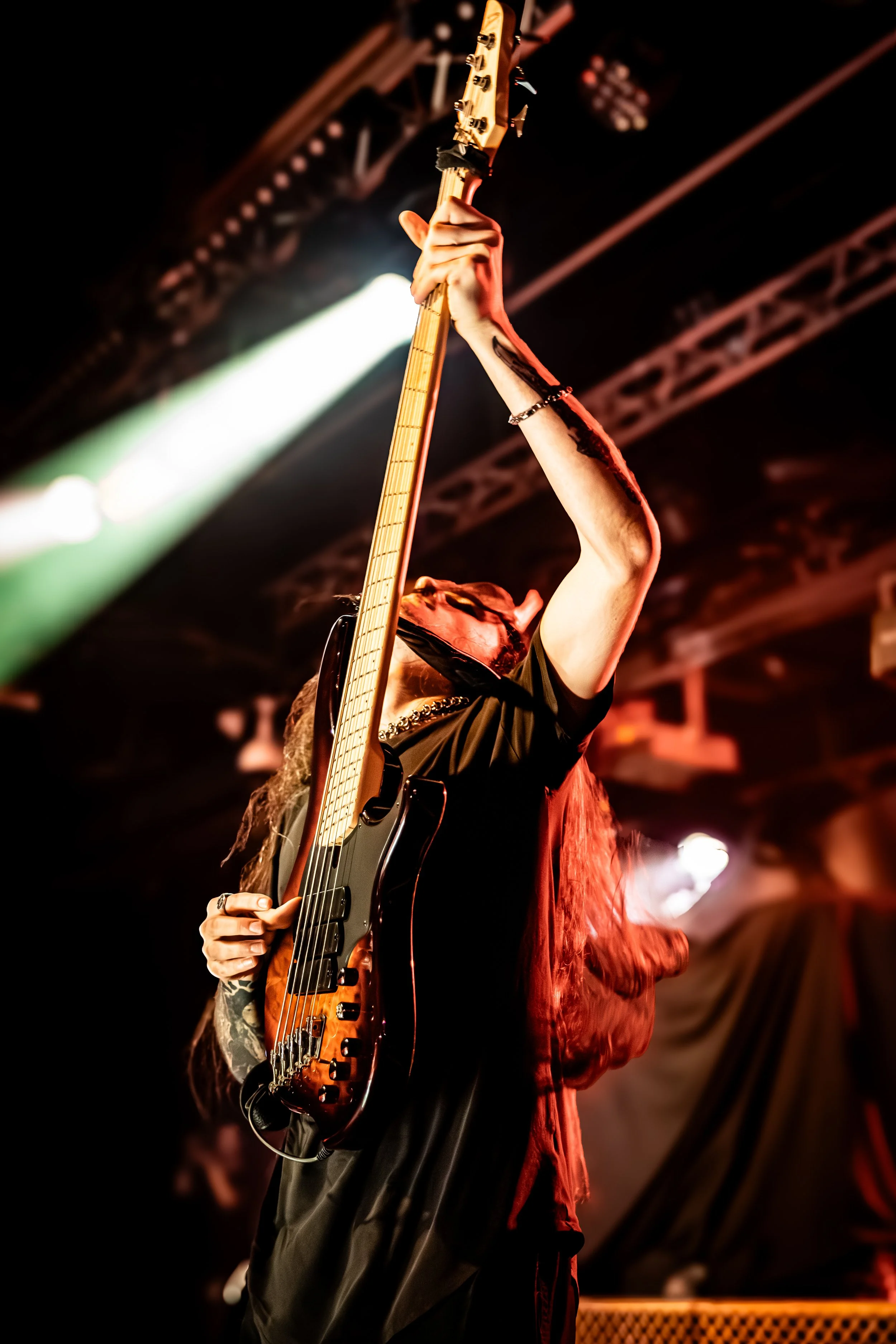 A musician holding an electric guitar with the neck raised above their head in a performance setting, with stage lighting and dark background.