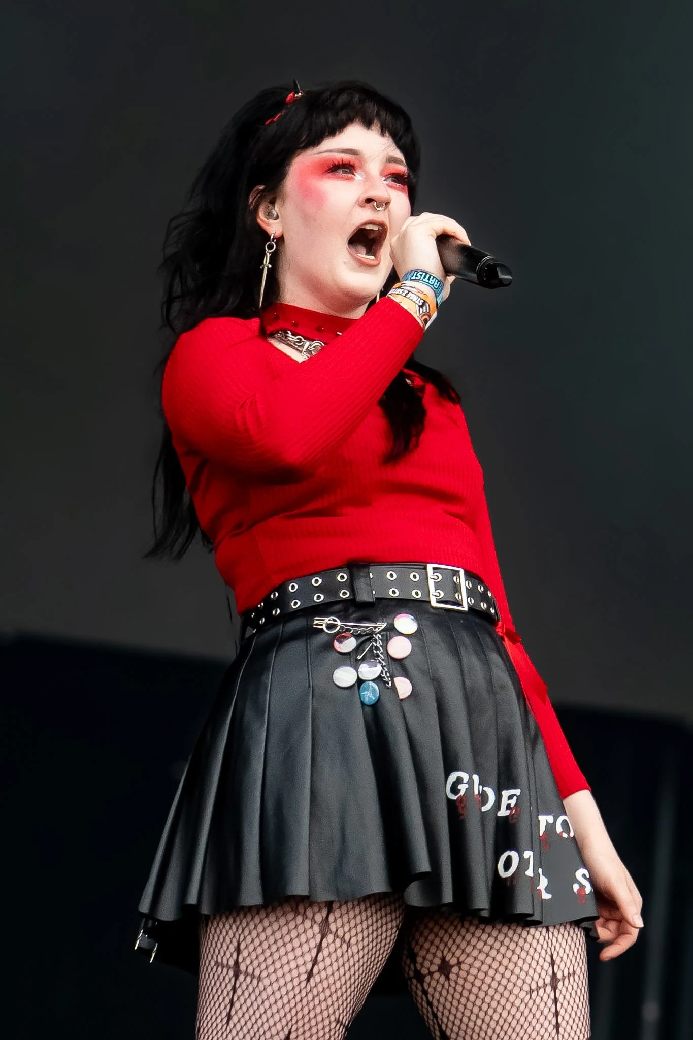 A woman singing into a microphone on stage, wearing a red top, black leather skirt decorated with patches, fishnet stockings, and jewelry with dark hair and makeup.