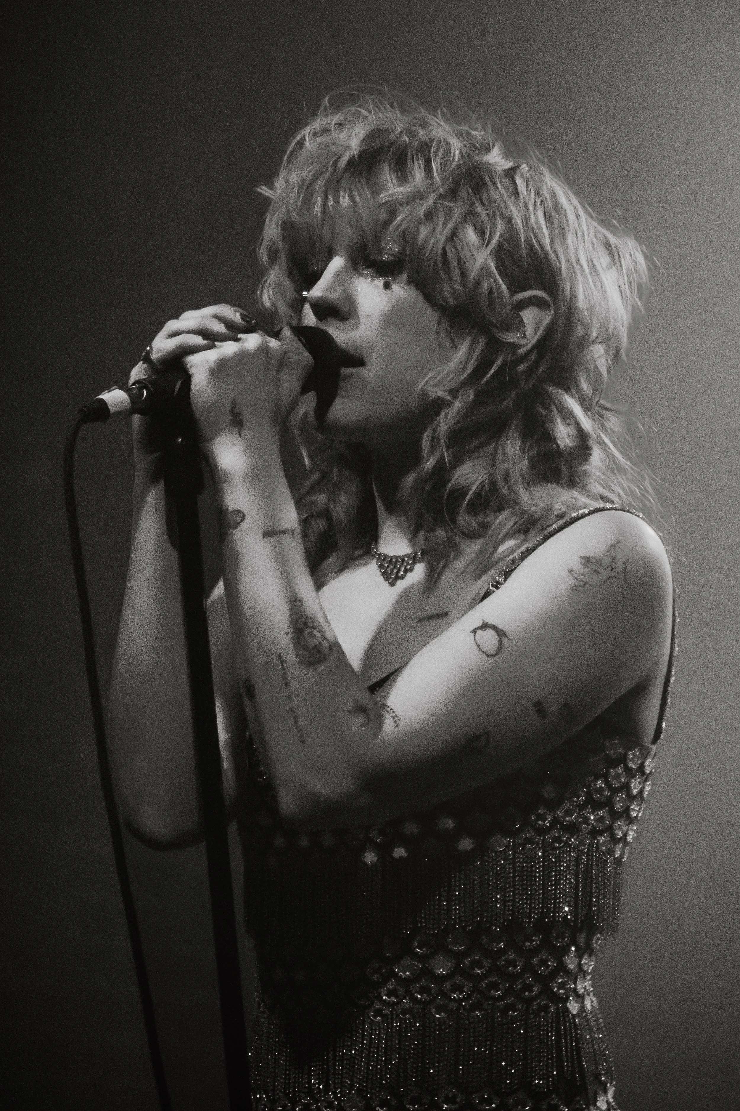 A woman with curly hair singing into a microphone, wearing a sparkly dress and jewelry, with tattoos on her arms, against a dark background.