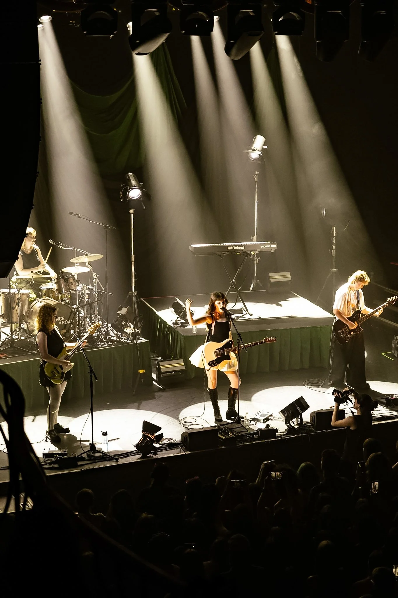 A live music performance on stage with multiple musicians, including a drummer, guitarists, and a singer, under stage lights in front of an audience.