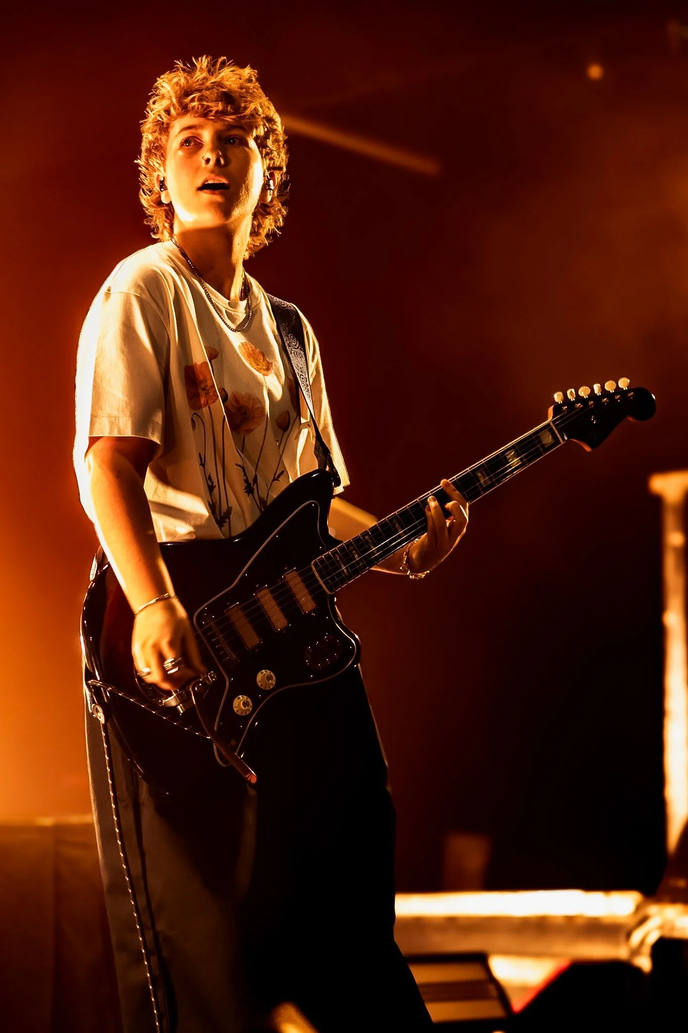 A young person with curly red hair playing an electric guitar on stage, illuminated by warm orange lighting.
