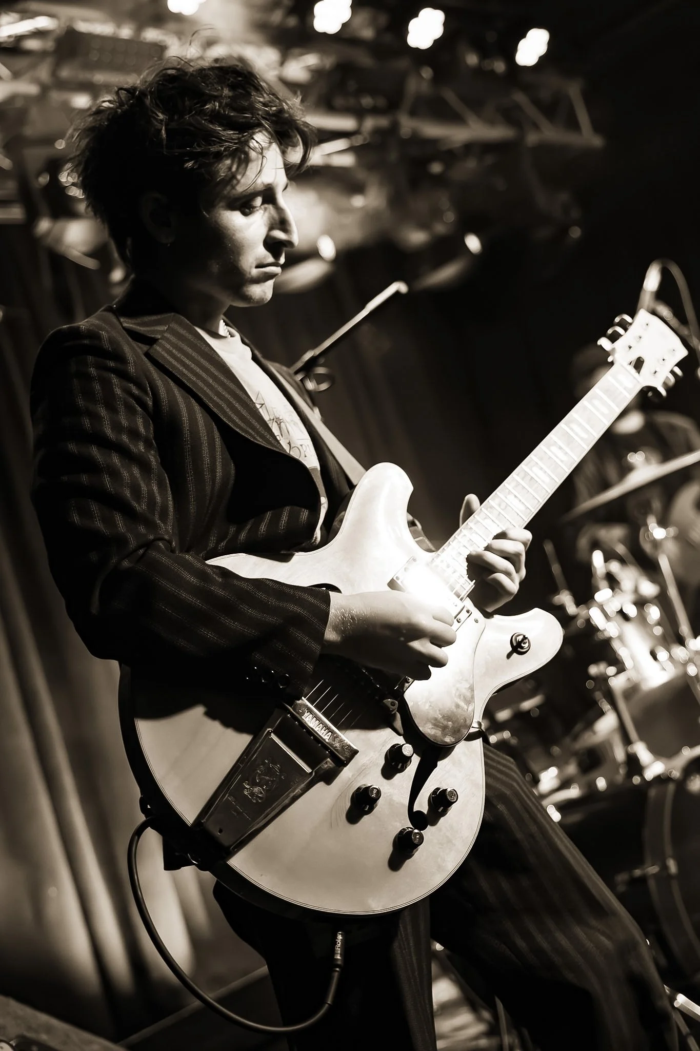 A person with short hair playing an electric guitar on stage, with a blurred background of instruments.