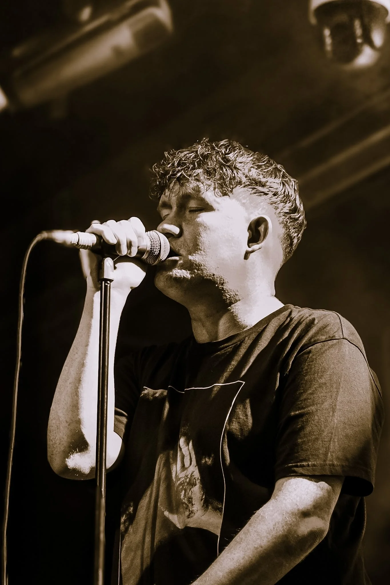 A young man with curly hair singing into a microphone, wearing a dark t-shirt.