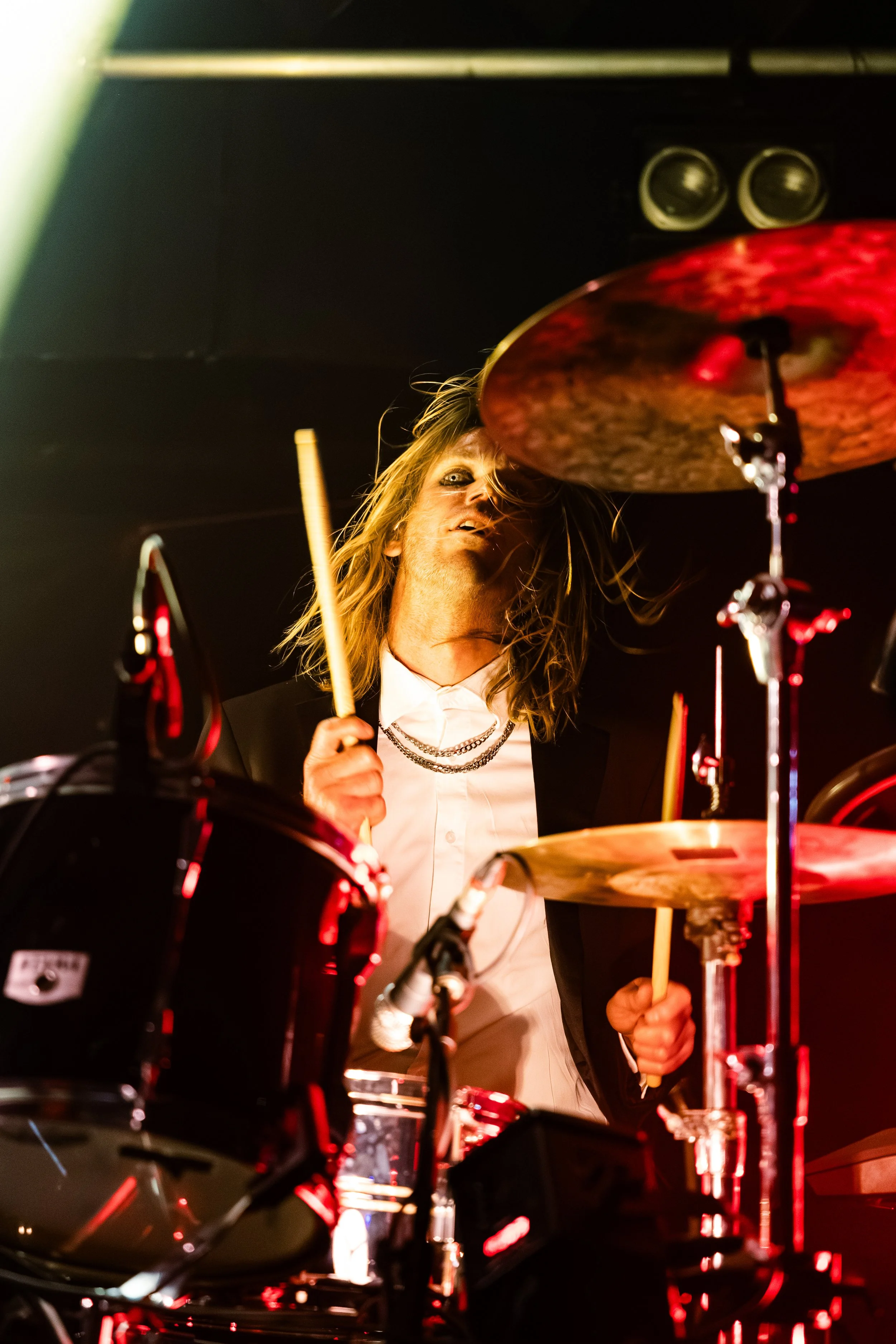 A male drummer with long hair, wearing a white shirt and black blazer, playing a drum set during a live performance, illuminated by stage lighting.