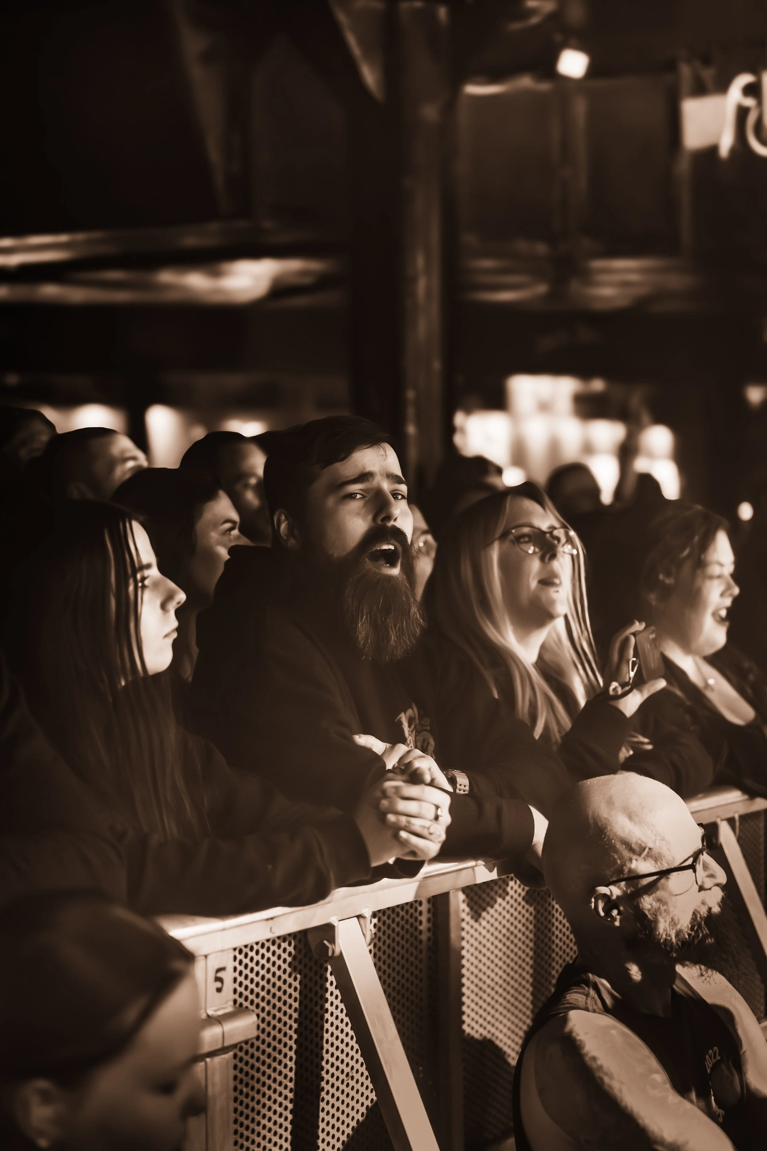 People sitting in a dark indoor venue, attentively watching a performance or event. Some are taking photos or videos with their phones.