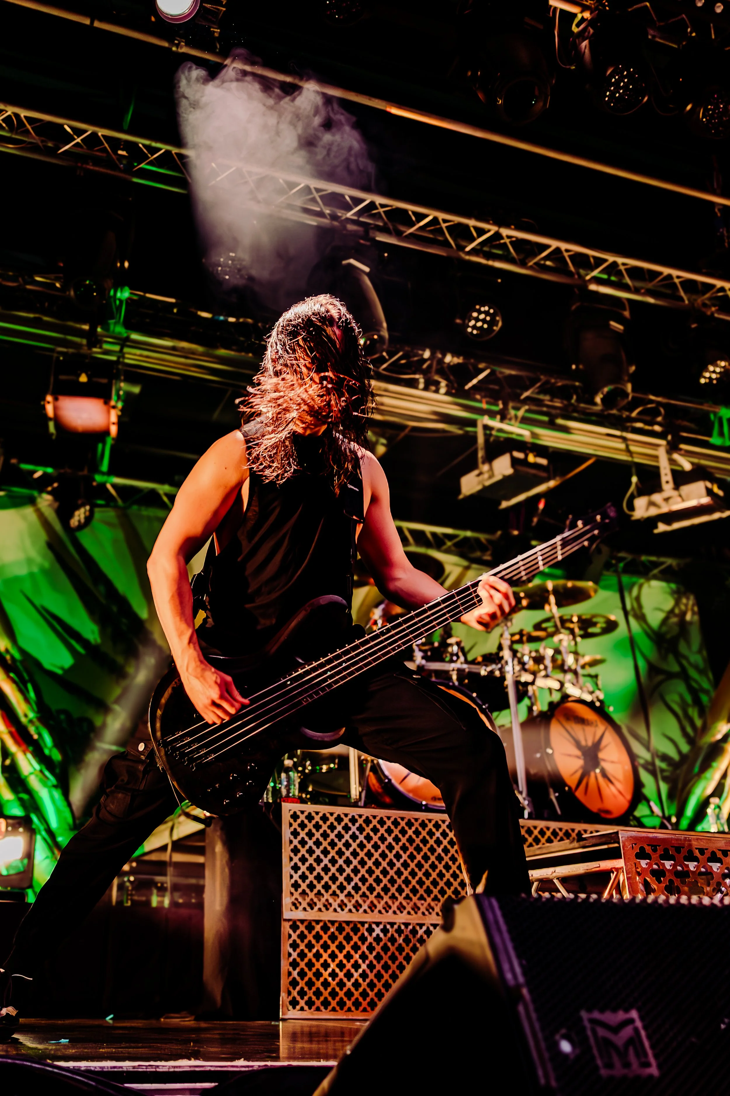 A musician playing a black electric bass guitar on stage during a live concert, with hair flying and stage lights overhead.