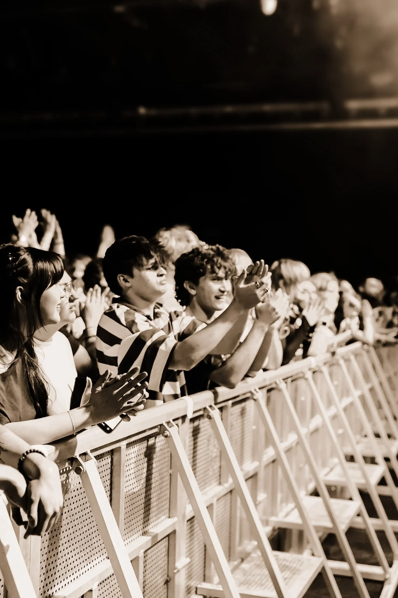 A group of people standing behind a metal barrier, clapping and smiling at an event, with some raising their hands in excitement.