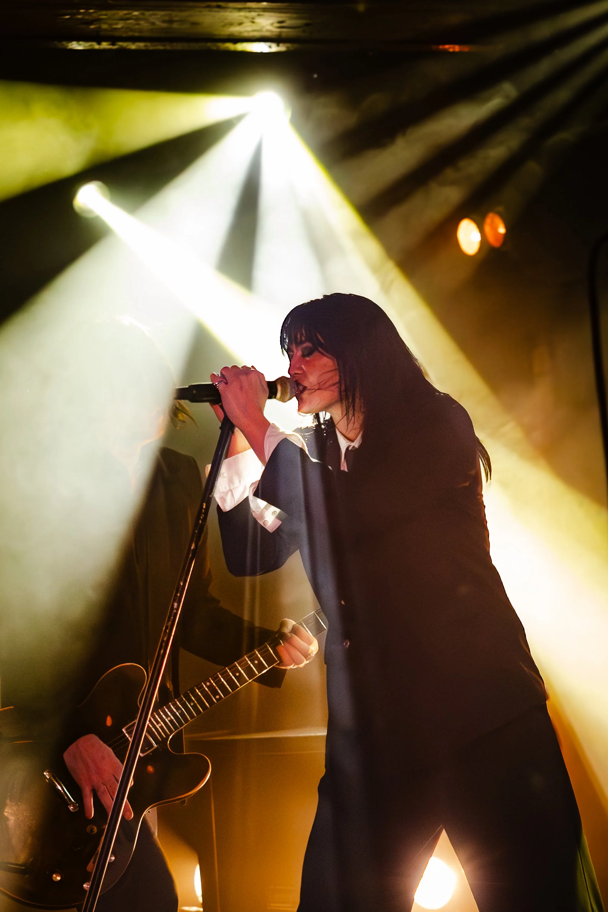 A female singer with long dark hair singing into a microphone, backlit by yellow stage lights, holding a small object in her hand, with another person playing an guitar beside her, in a dimly lit concert setting.