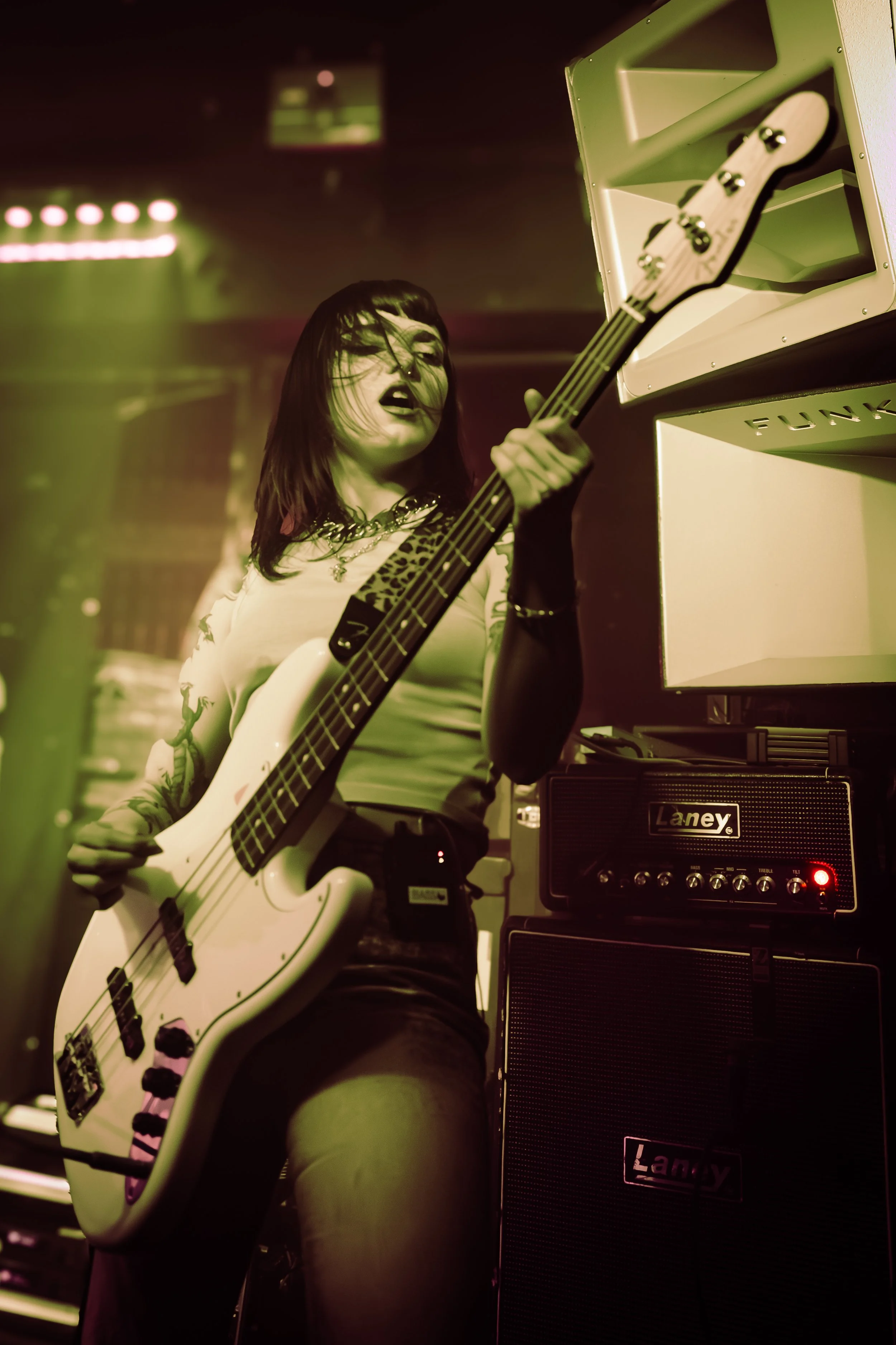 Female musician playing a white bass guitar on stage with a guitar amplifier nearby.