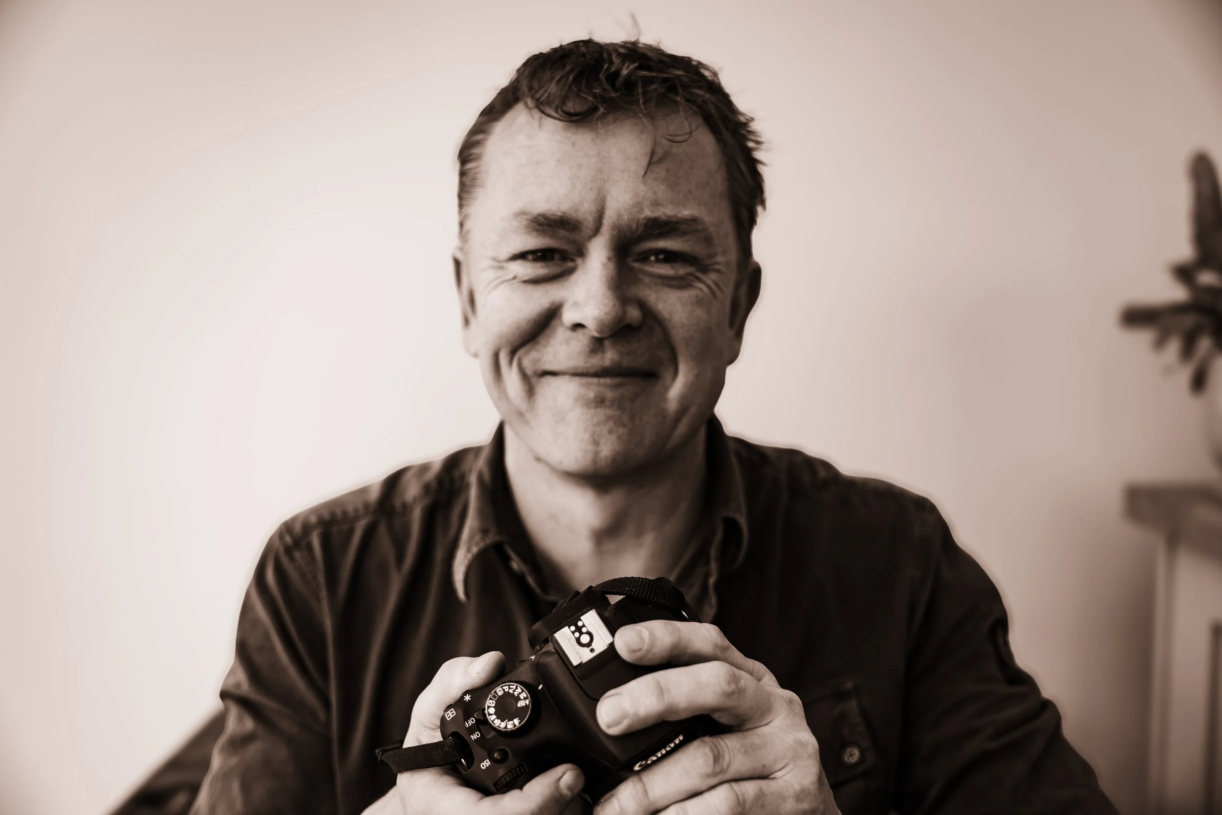A smiling man with short curly hair holding a camera in his hands, wearing a dark shirt, standing in front of a plain wall.