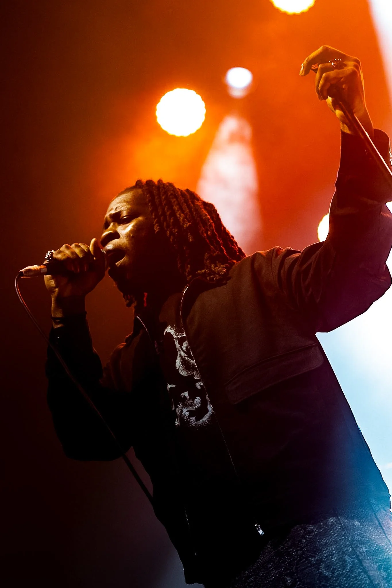 A performer singing into a microphone on stage with dramatic orange and dark lighting.