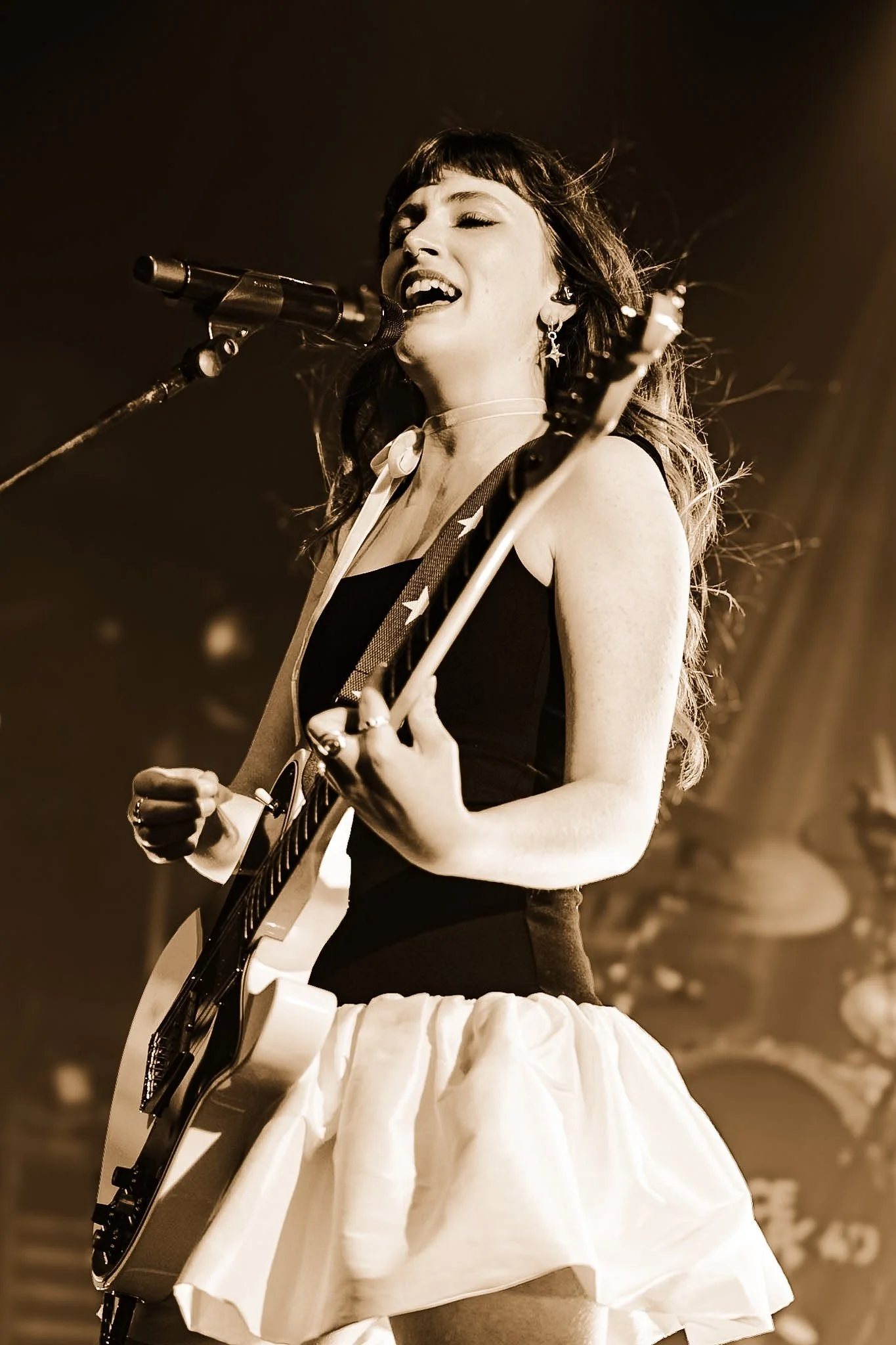A female singer passionately performing on stage, holding a guitar, with her eyes closed and mouth open, wearing star-shaped earrings, a black top, and a white skirt.