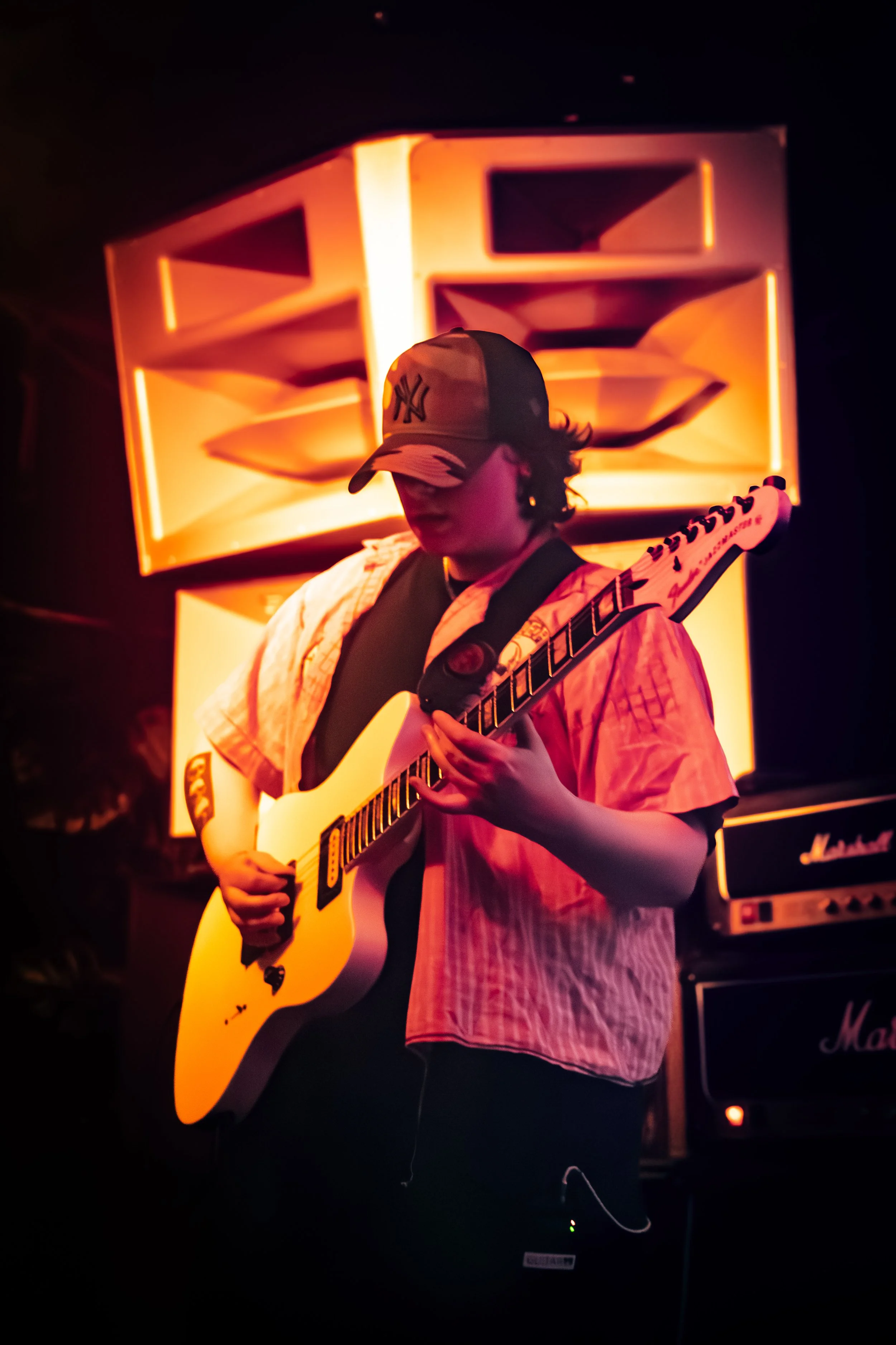 Person wearing a cap and headphones playing an electric guitar in a dimly lit setting with an illuminated brick wall and Marshall amplifier in the background.