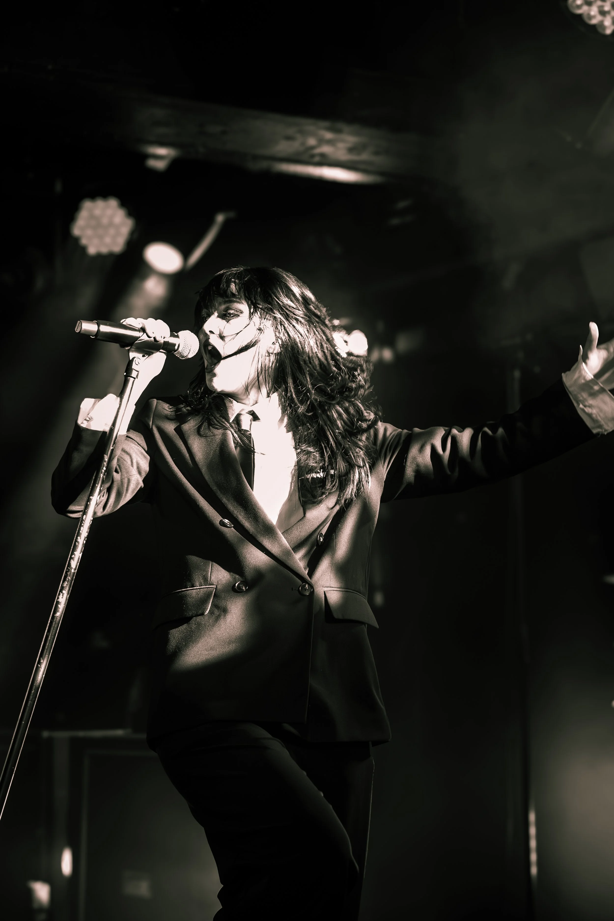 A woman singing passionately on stage with a microphone, wearing a blazer and gesturing with one arm extended, in a dark concert setting with stage lights.