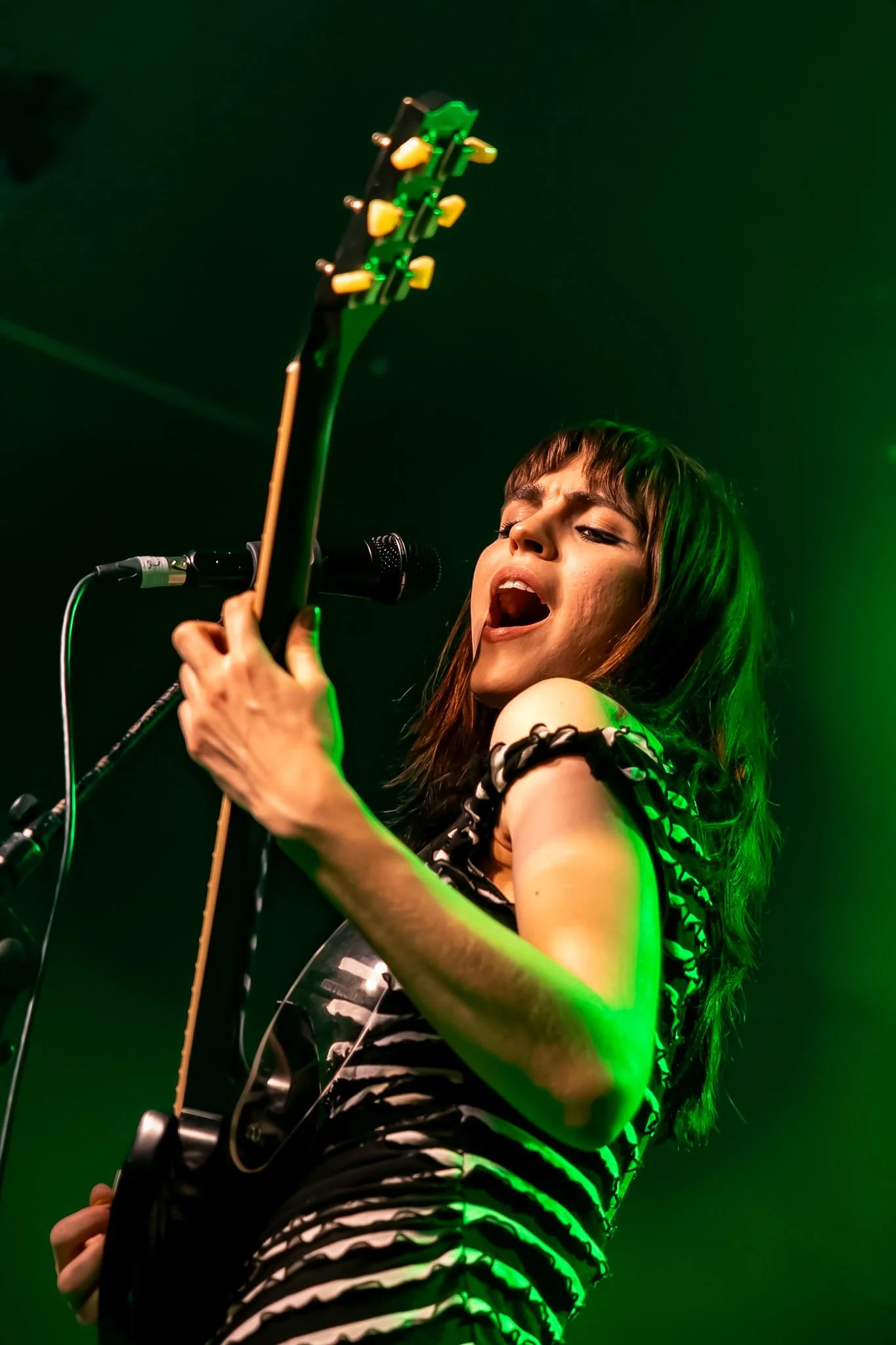 A female musician passionately singing into a microphone while playing an electric guitar, wearing a black and white striped outfit, with green stage lighting