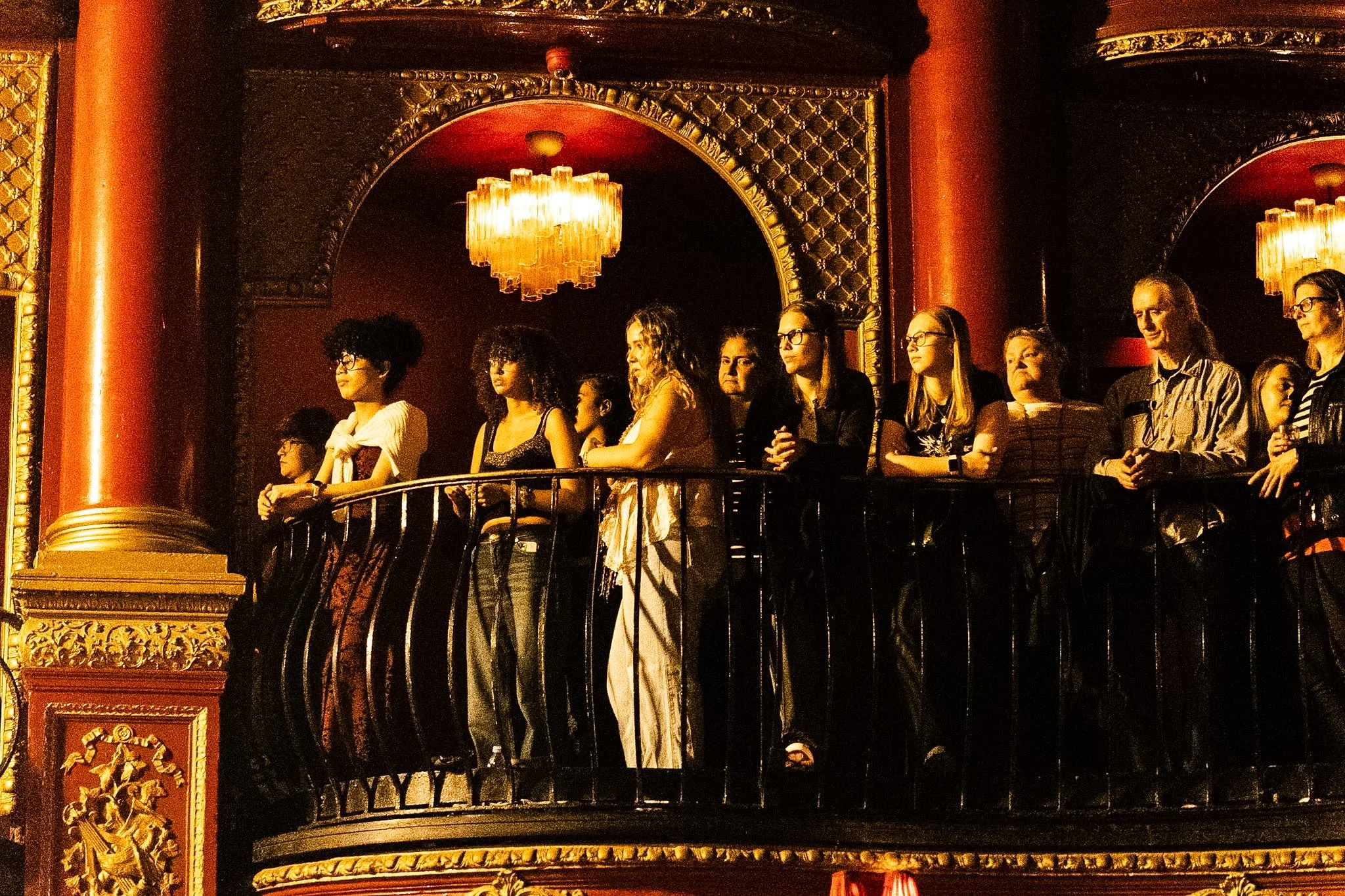 People standing on a balcony inside a historic theater, with ornate gold decorations and a chandelier hanging from the ceiling.