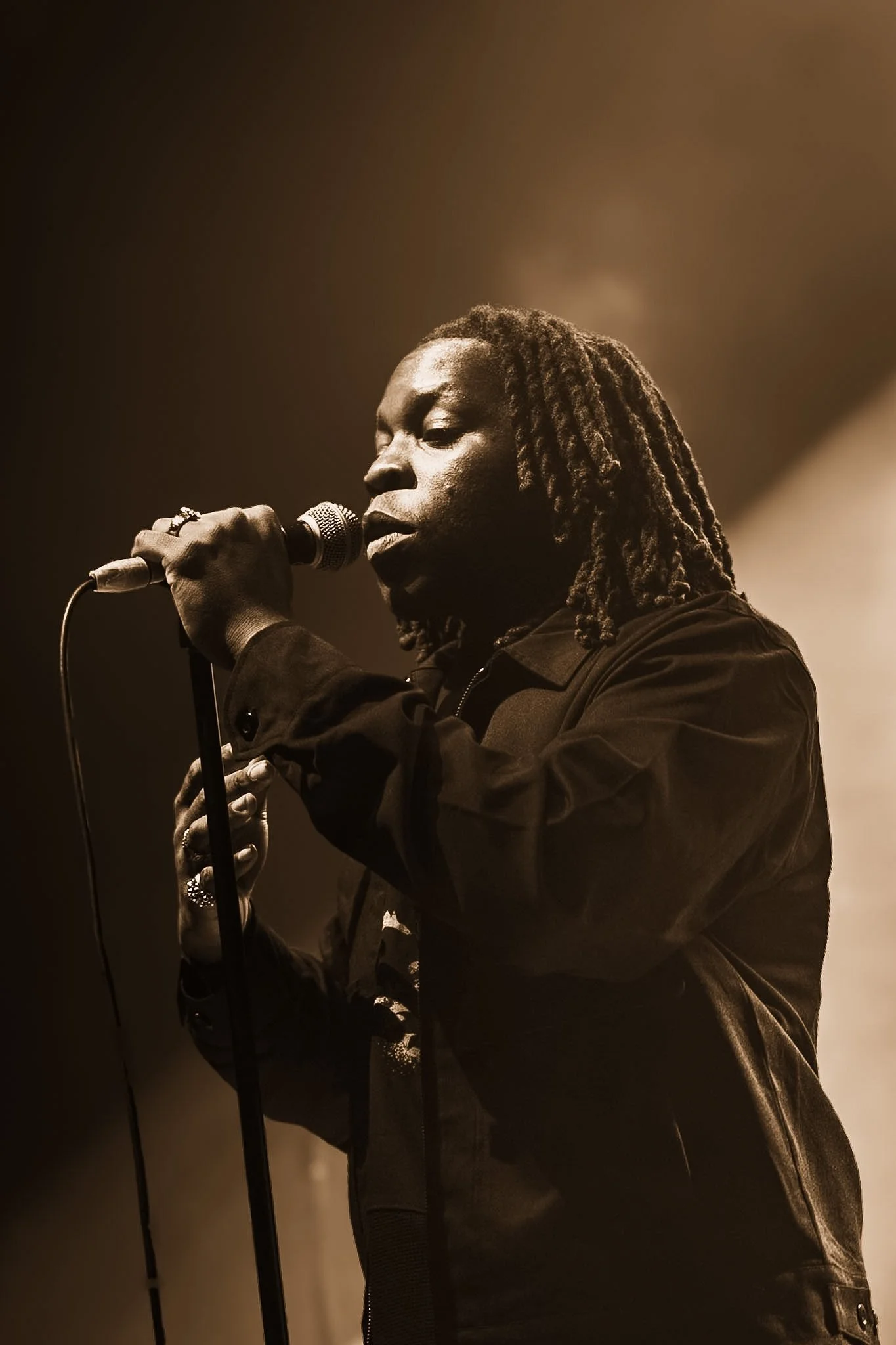 A person with dreadlocks singing into a microphone on a dimly lit stage.