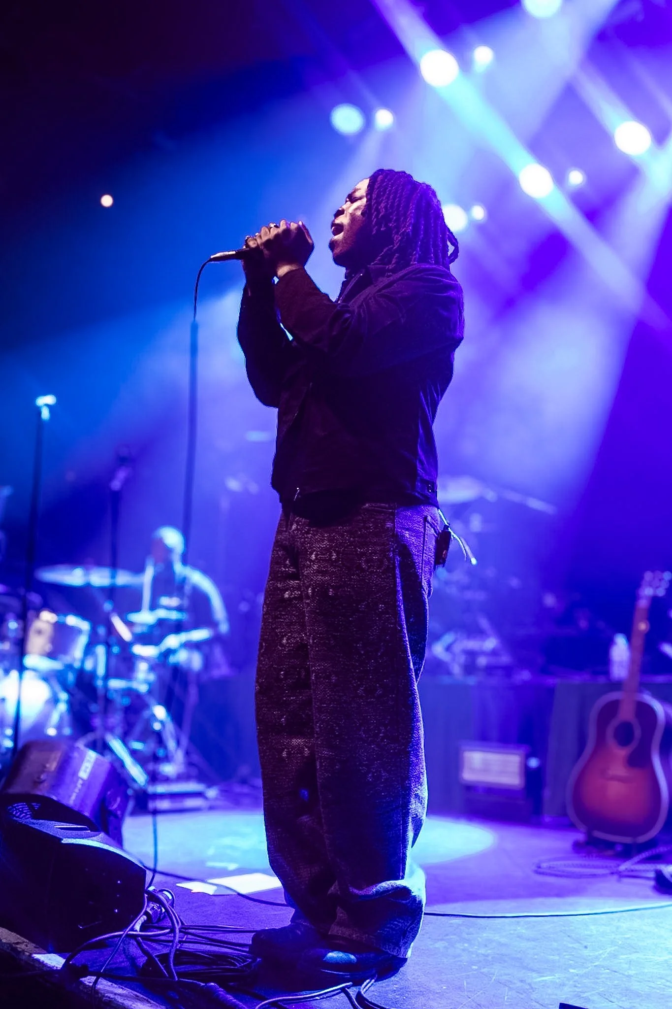 A singer with long dreadlocks performs on stage, holding a microphone with stage lights illuminating the background.
