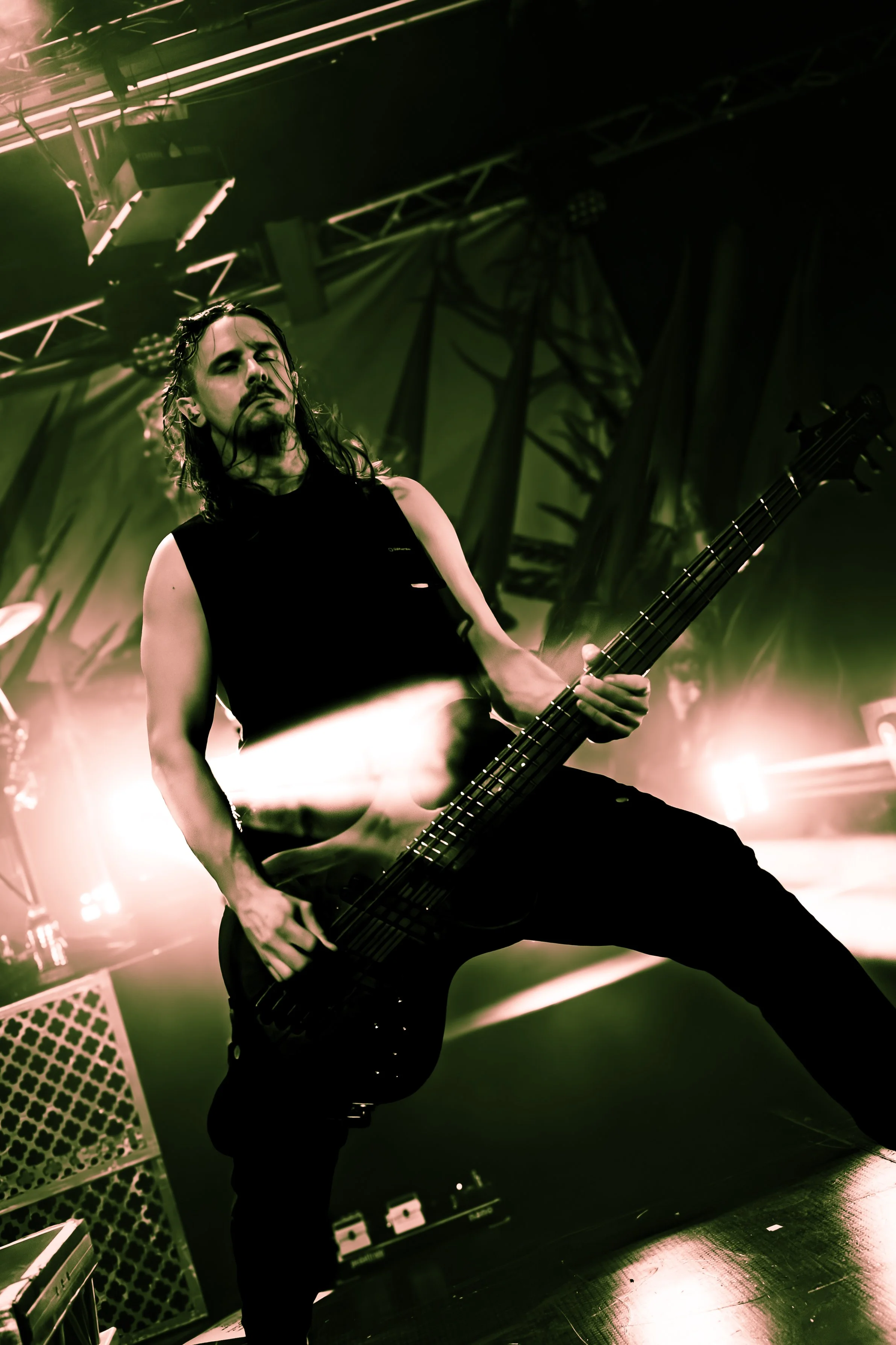 A musician with long hair and a beard playing an electric bass guitar on stage under green lighting, wearing a sleeveless black shirt, in a confident pose with one leg forward.