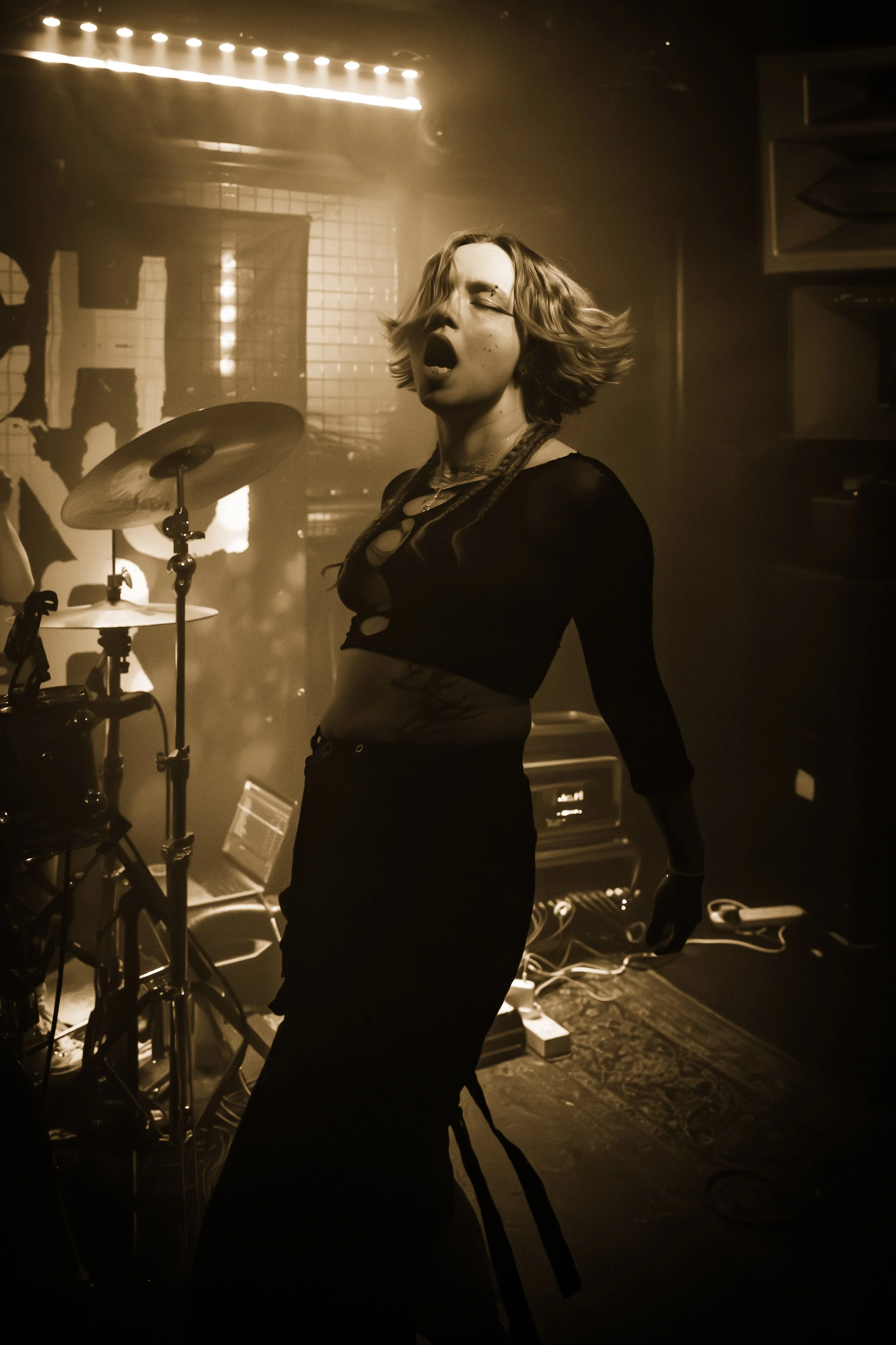 A woman with short, wavy hair singing passionately on stage with her eyes closed, surrounded by musical equipment and dim, moody lighting.