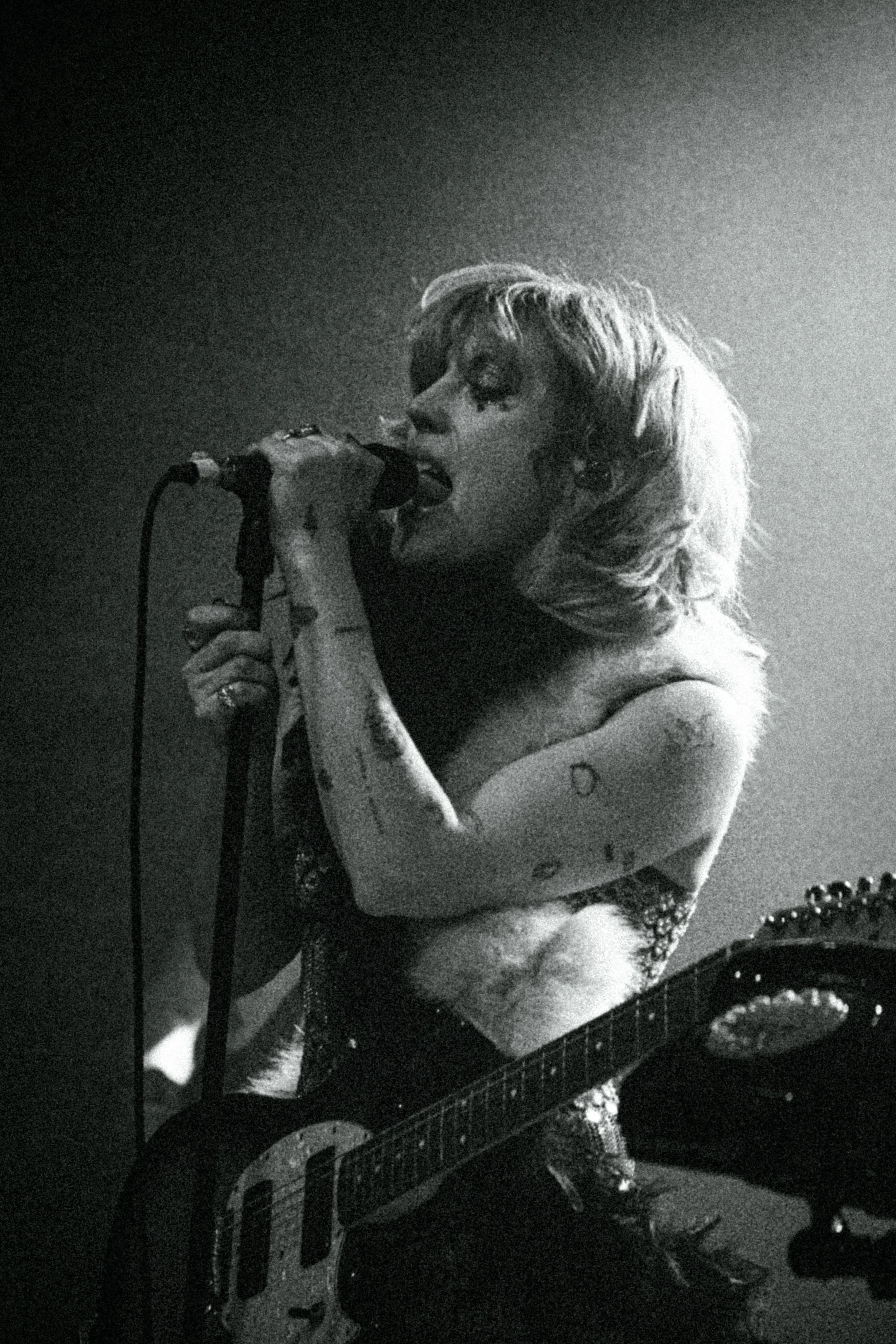 A black and white photo of a female singer with short, wavy hair, singing into a microphone while playing an electric guitar. She has tattoos on her arms and is wearing a sleeveless top with sequins.