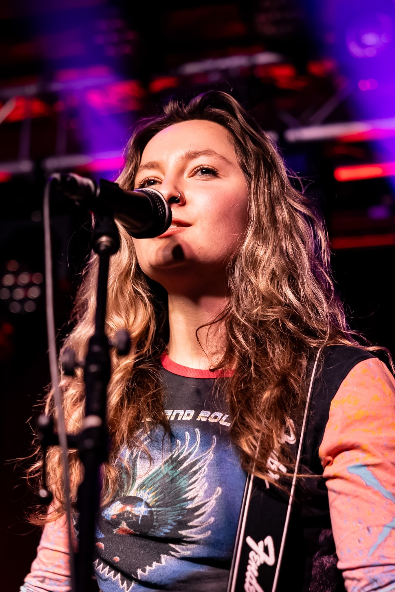 A woman with long wavy hair and a nose ring singing into a microphone on stage, with colorful purple and pink lights in the background.