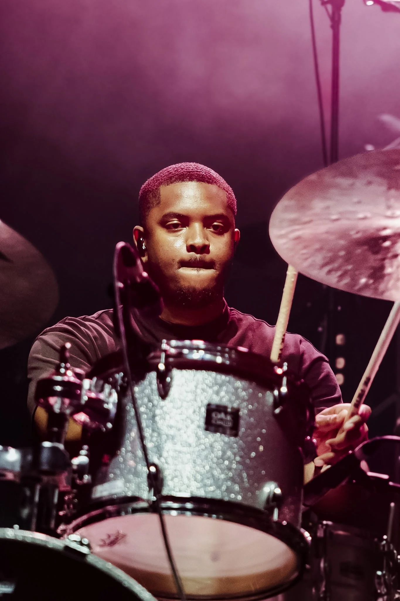 A drummer with short hair, intense expression, wearing a black shirt, playing drums in a dimly lit setting with pink and purple lighting.