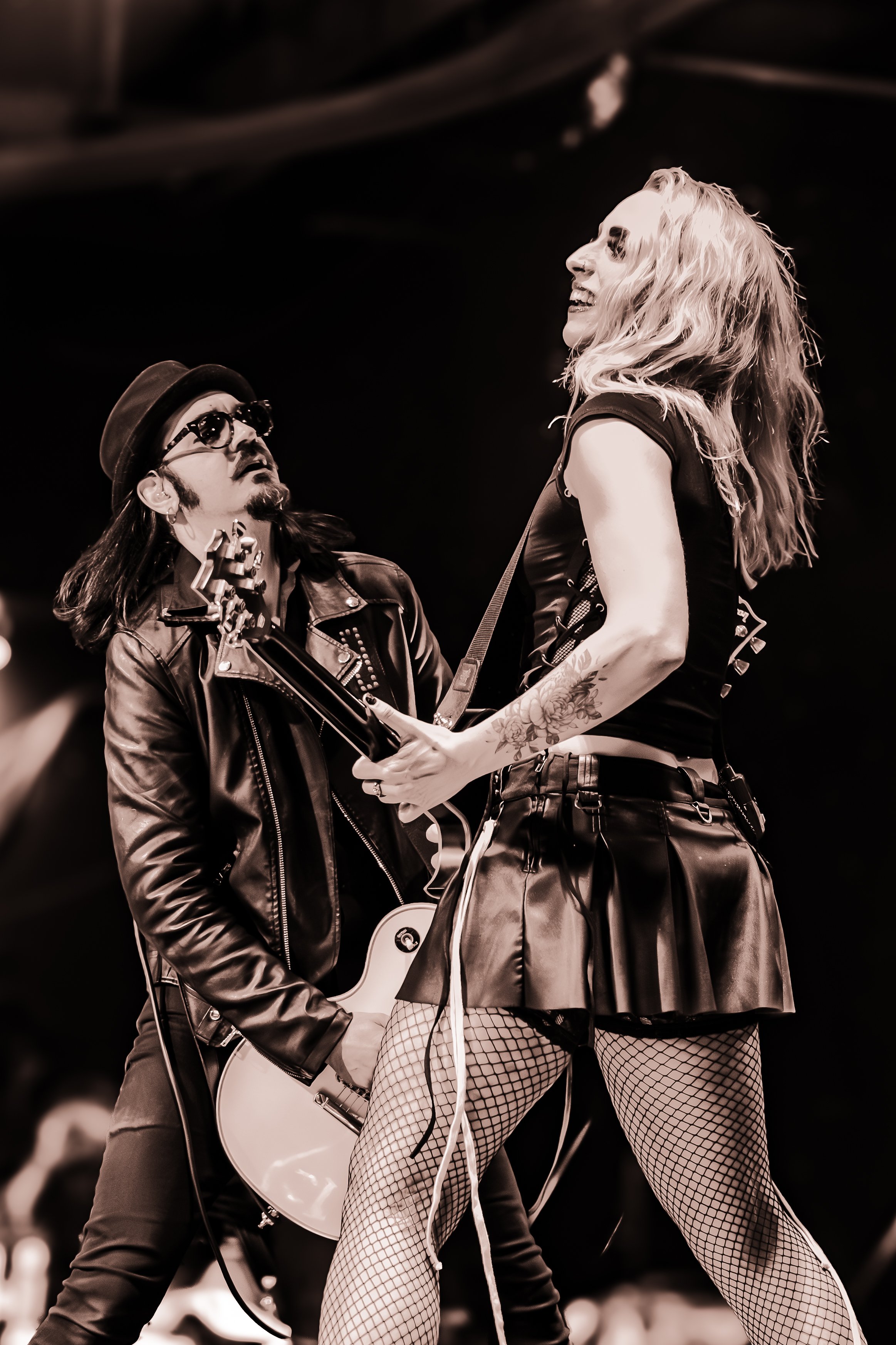A male guitarist wearing a leather jacket, glasses, and a fedora hat, looking up at a female singer with long, wavy hair, wearing a black sleeveless top, a pleated skirt, fishnet stockings, and tattoos on her arm, performing on stage.