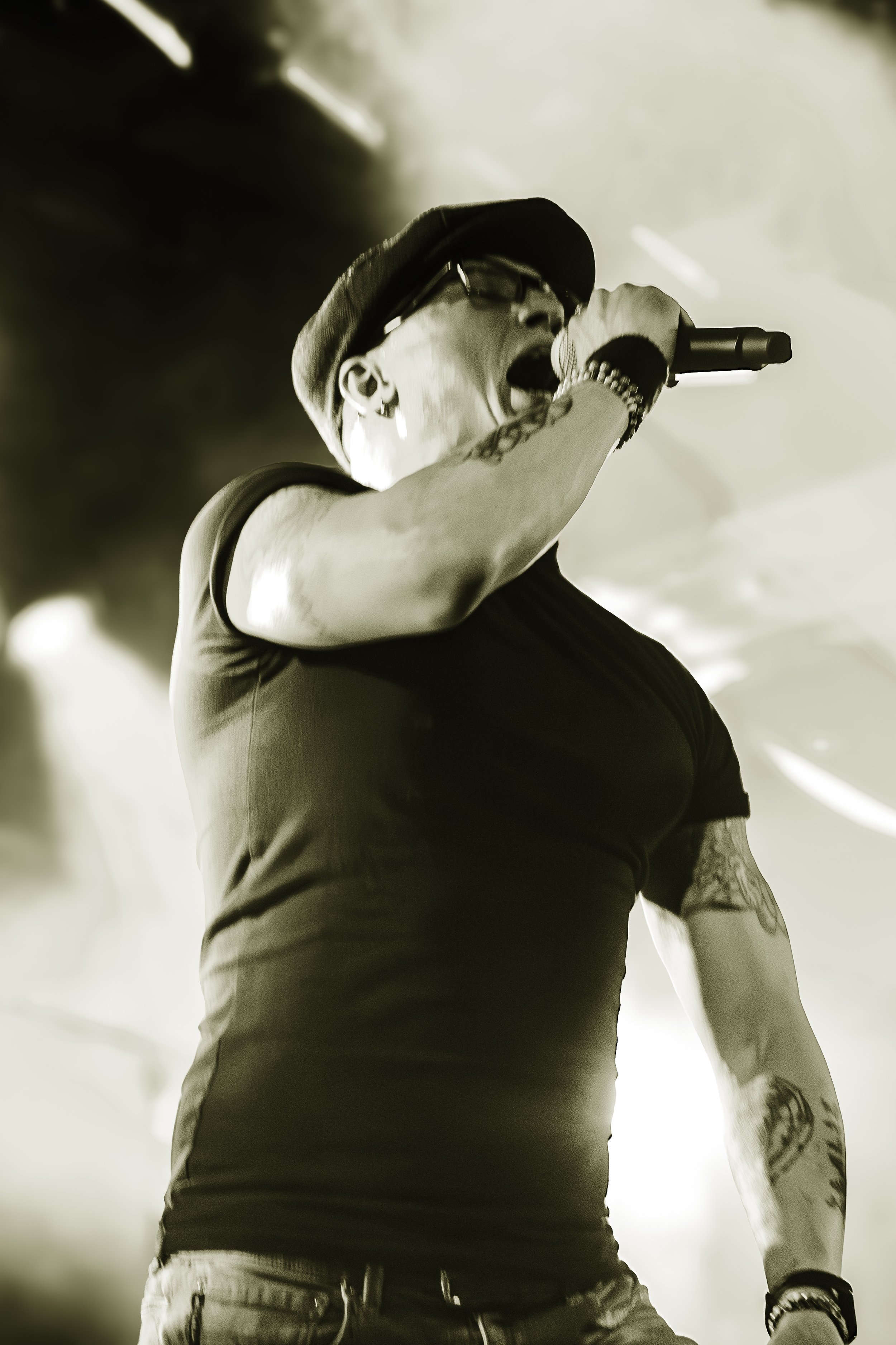 A black and white photo of a person singing into a microphone, wearing glasses, a cap, and a t-shirt.