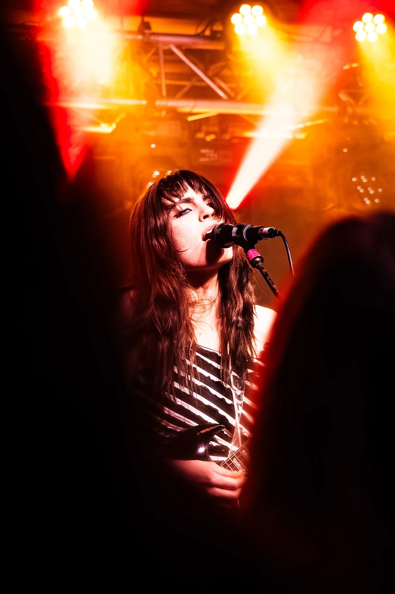 A female singer with long dark hair and bangs performing on stage with a microphone, seen in front of bright red and yellow stage lighting, holding a guitar, and surrounded by a dark audience.