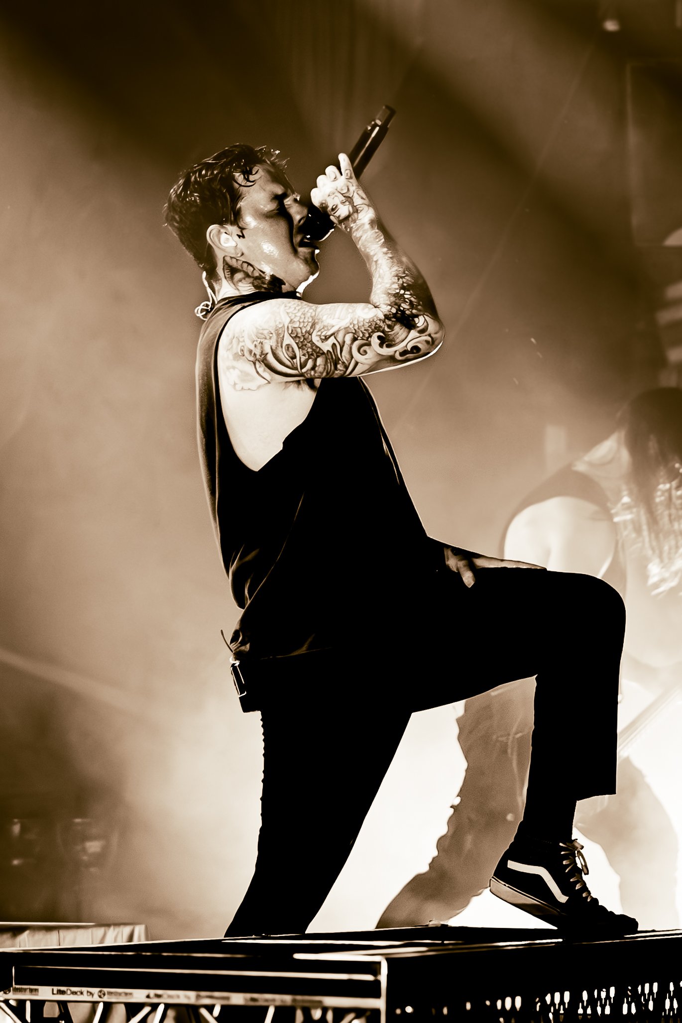 A performer singing into a microphone on stage, with tattoos on his arm, wearing a sleeveless shirt and sneakers. The image is in black and white.