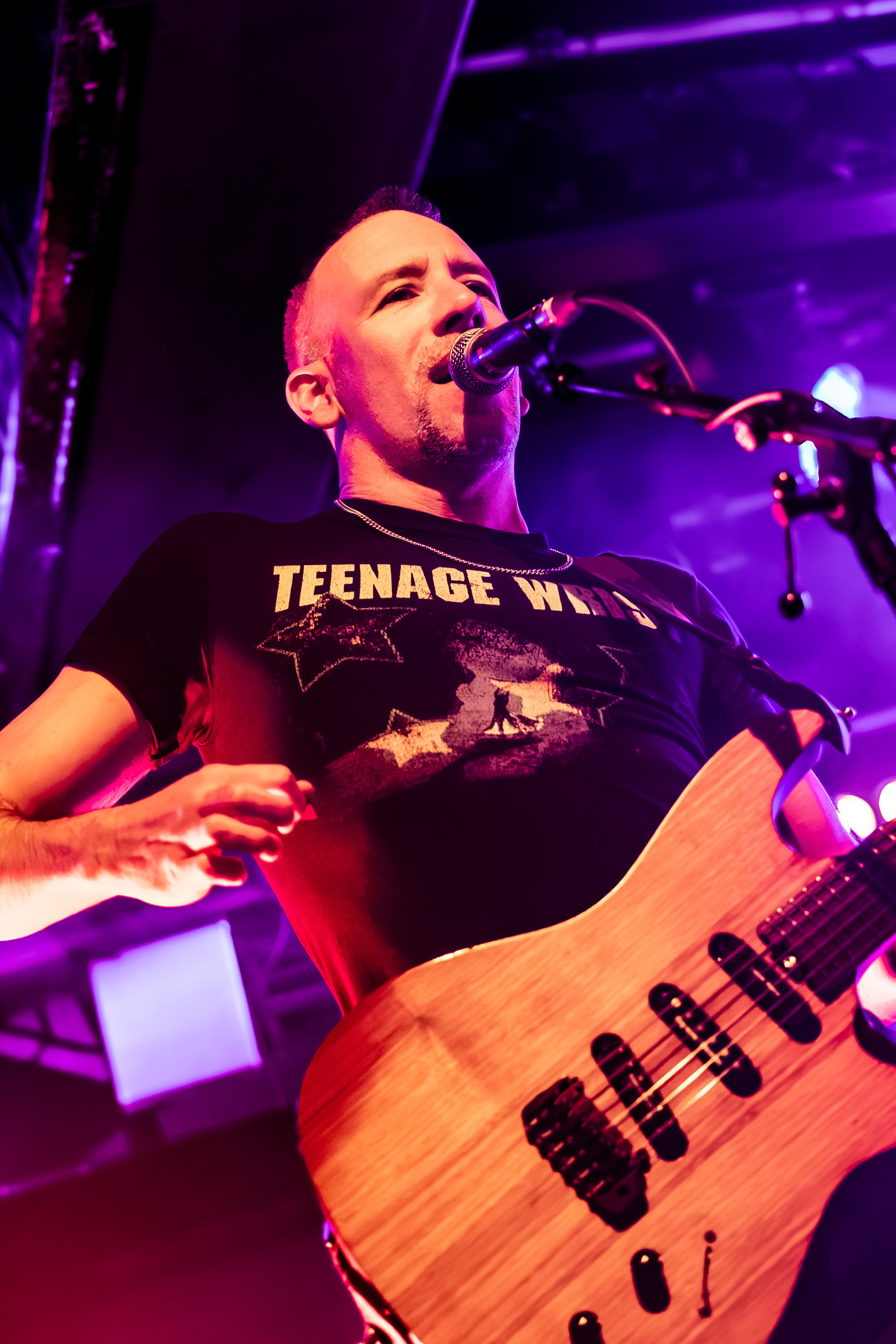 A male musician with a Mohawk hairstyle singing into a microphone while playing an acoustic guitar on stage illuminated with purple and pink stage lights.