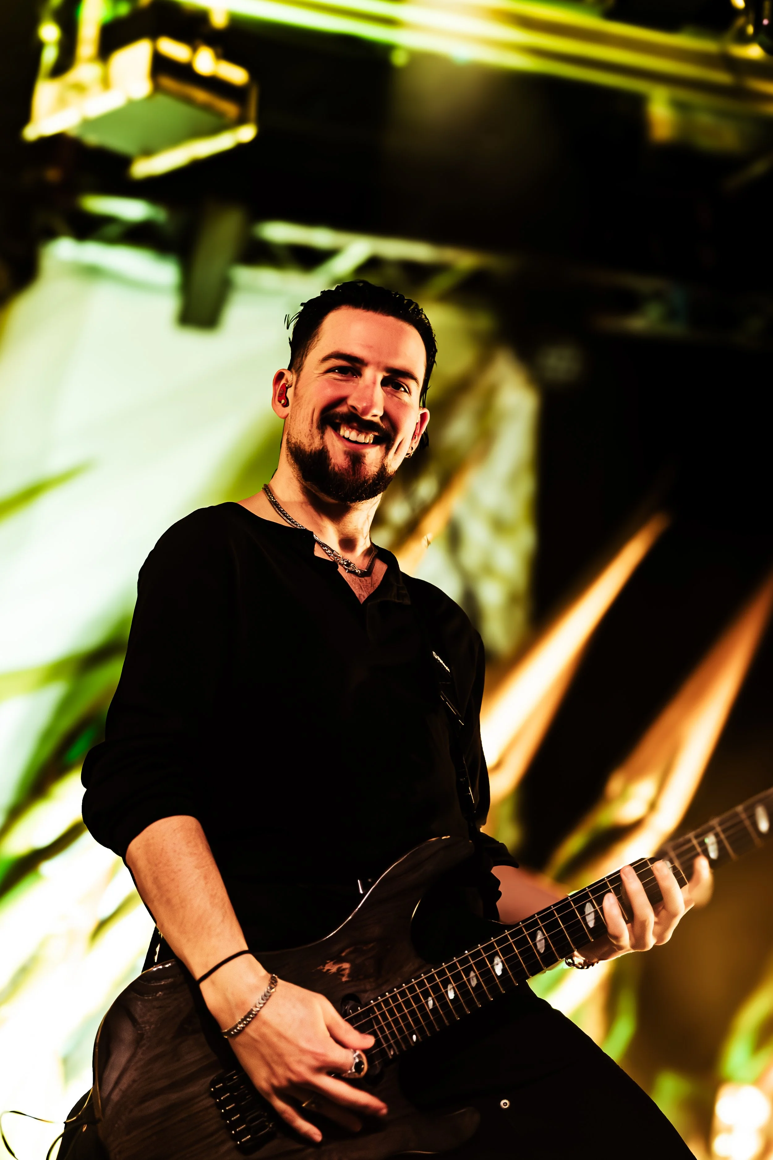 A man with dark hair and a beard playing an electric guitar on stage, smiling, with a blurred colorful background.
