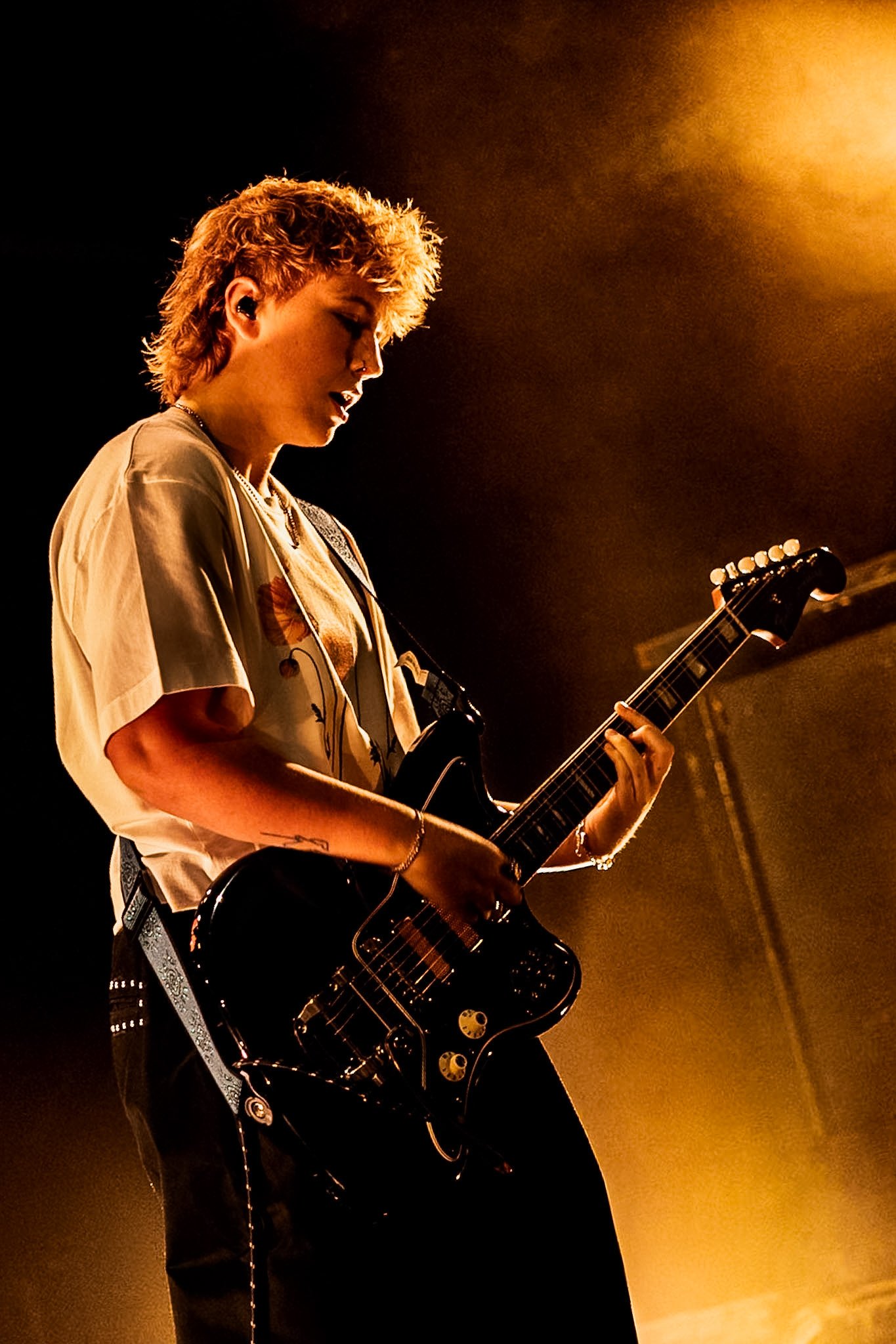 A person playing an electric guitar on stage under warm lighting, with short curly hair and a casual shirt.