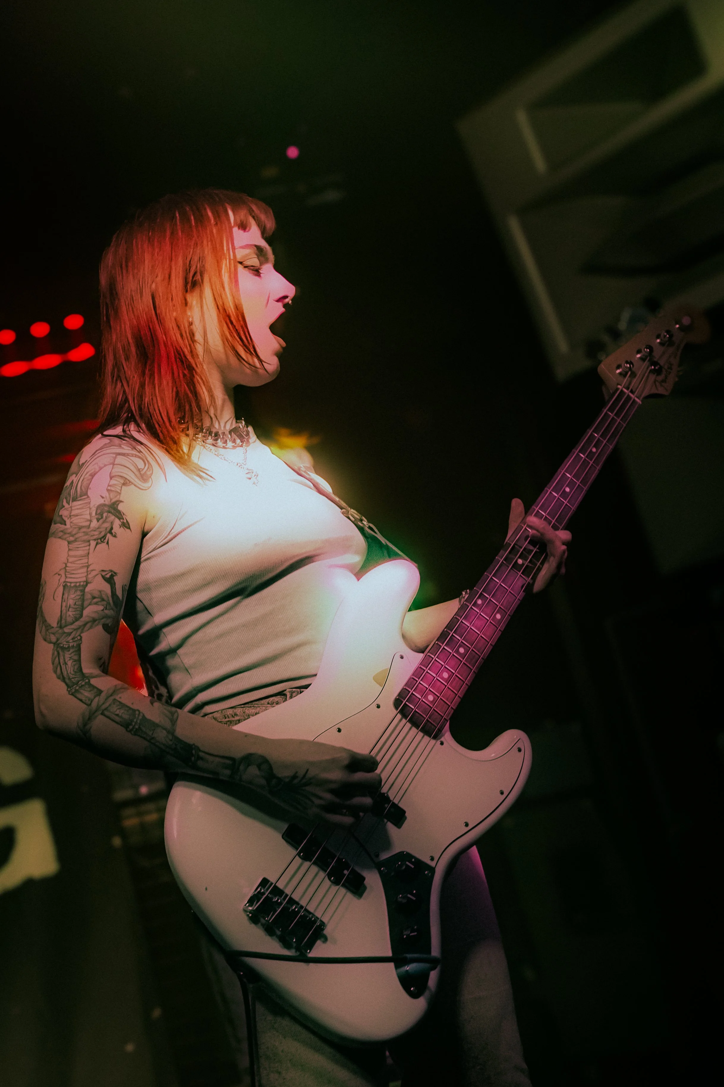 A woman with red hair, tattoos on her arms, wearing a beige top, playing a white electric bass guitar on stage with dark background and red stage lights.