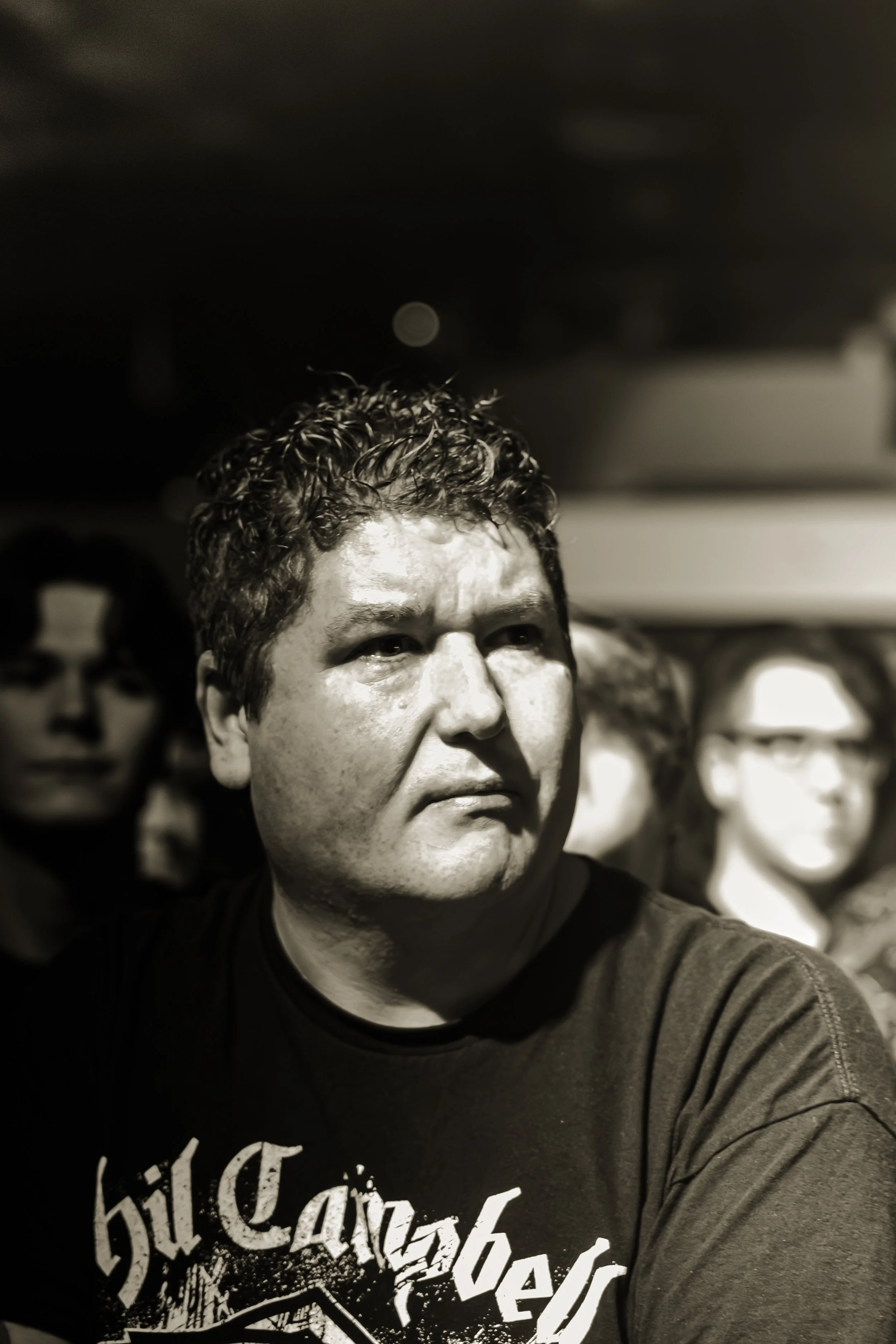 Closely cropped portrait of a middle-aged man with curly hair wearing a dark graphic T-shirt, with a blurred background.