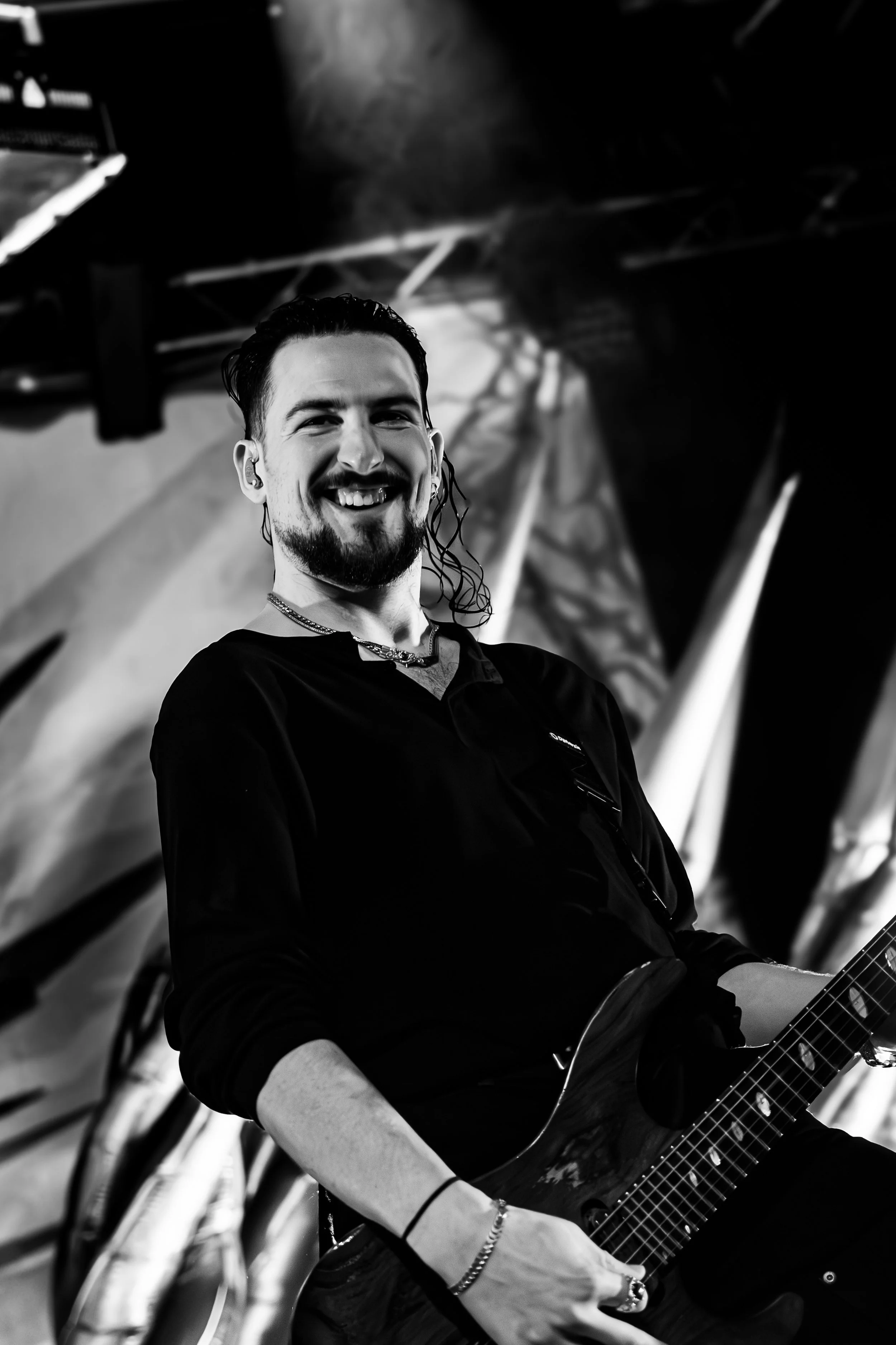 A young man with dark, wet hair, a beard, and a mustache smiling while holding a guitar, standing in front of a blurred background with structural elements.