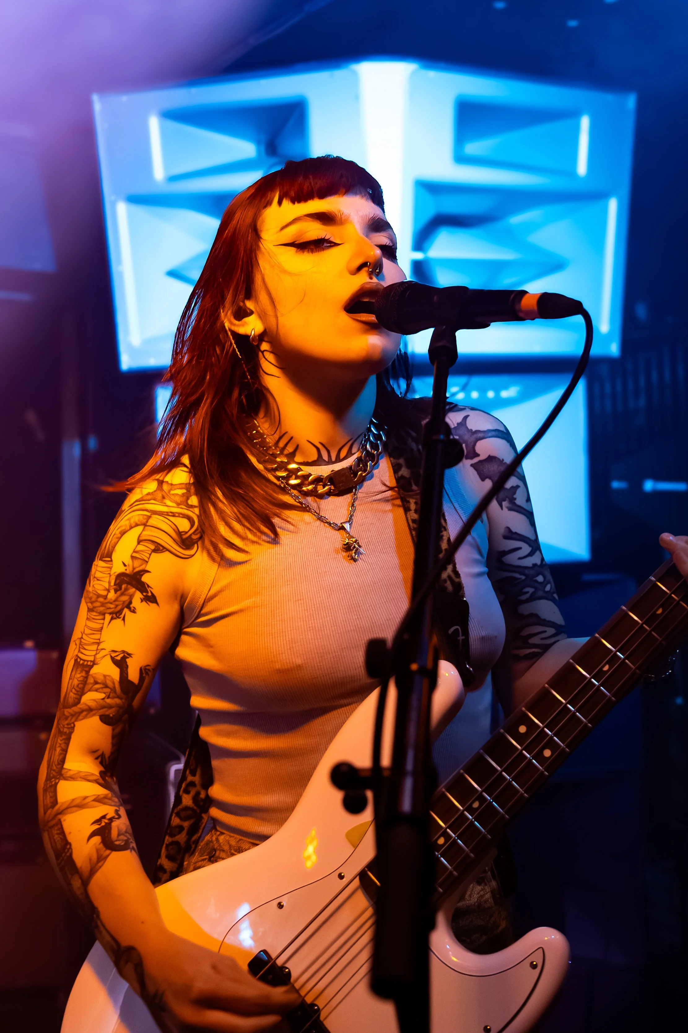 A female musician with tattoos and dark makeup singing into a microphone while playing an electric guitar, with a neon blue geometric light display in the background.