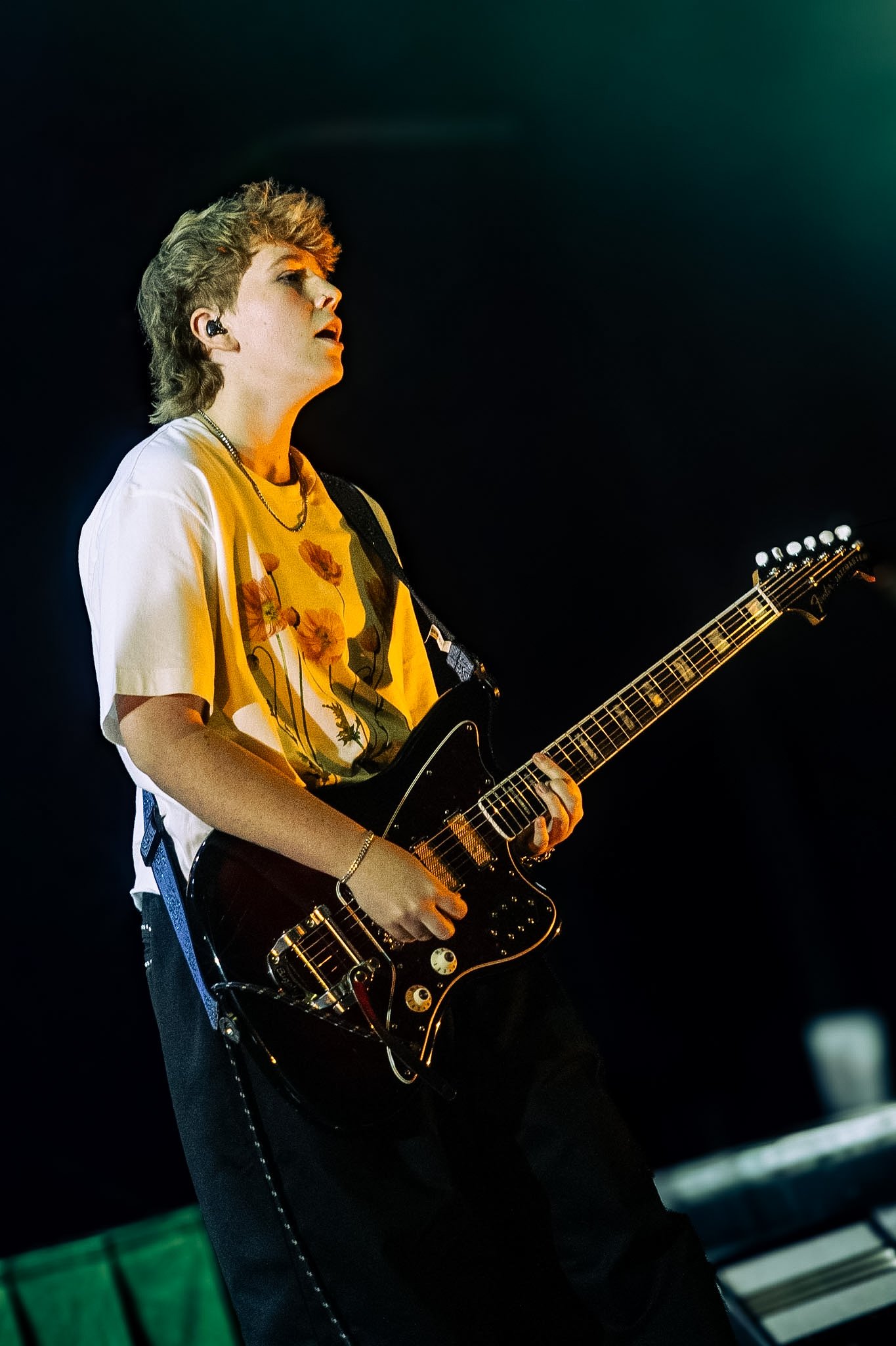 A young person with short, styled hair playing an electric guitar on stage, wearing a yellow shirt and black pants, with dark background lighting.