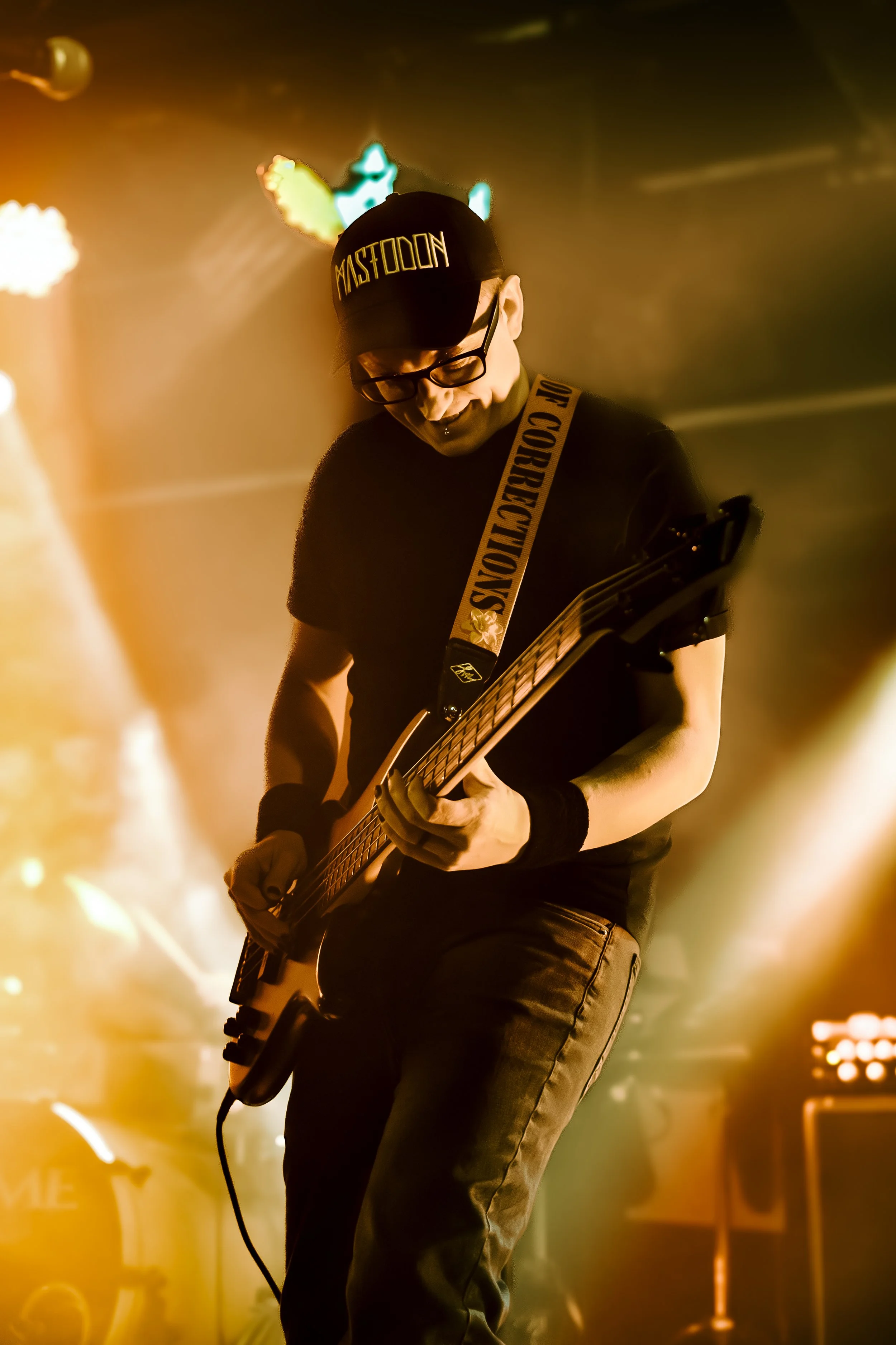 A person playing an electric guitar on stage, wearing a black cap with the word 'MASTODOON' and a black shirt. The stage is lit with warm, golden lights with a blurred background.