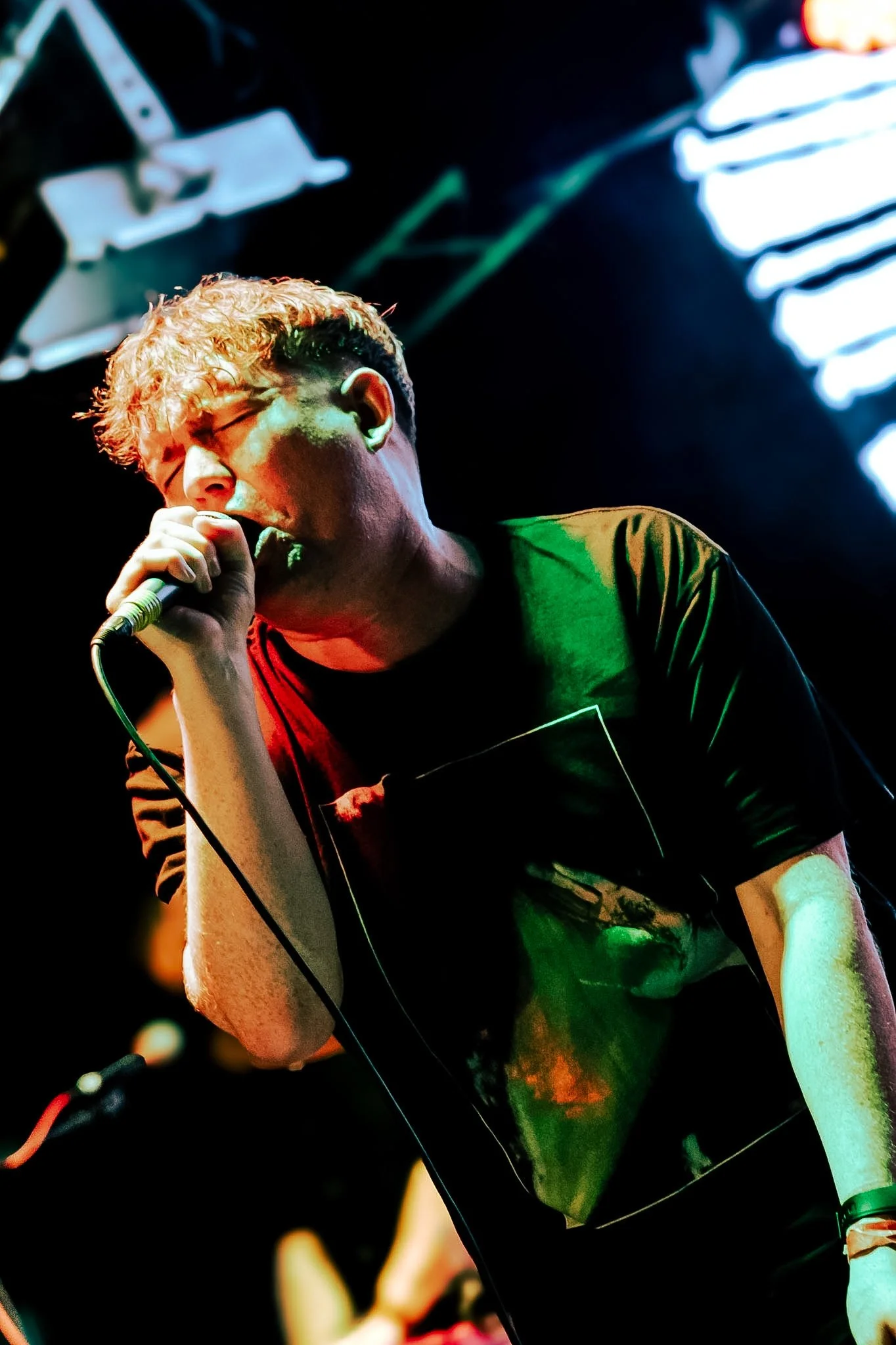 A young man with blond curly hair singing into a microphone on stage with vibrant lighting.