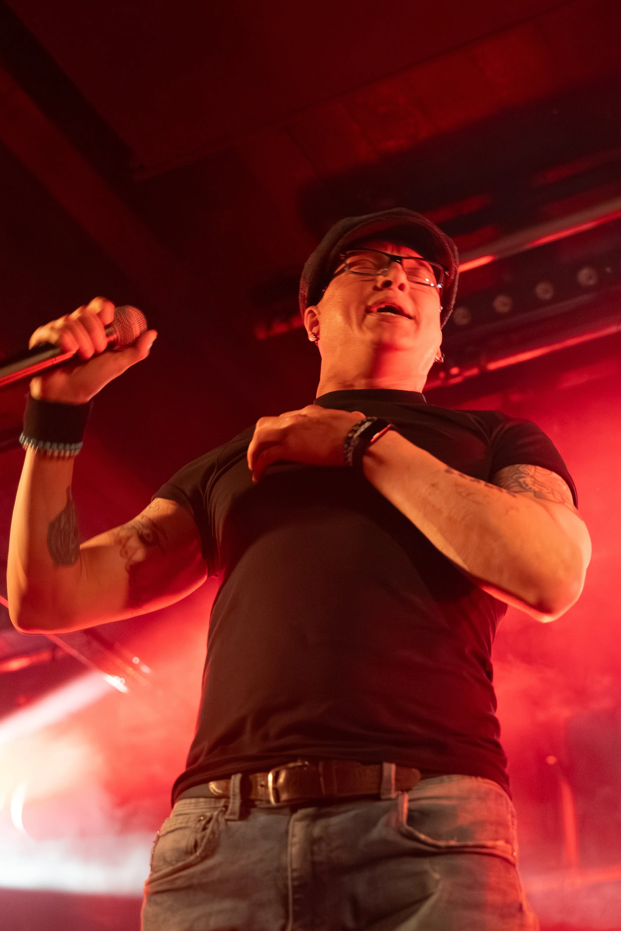 A person holding a microphone, wearing glasses, a hat, and a black t-shirt, standing in a stage or club with red lighting. A hand with a tattoo is visible, touching the person's shoulder.