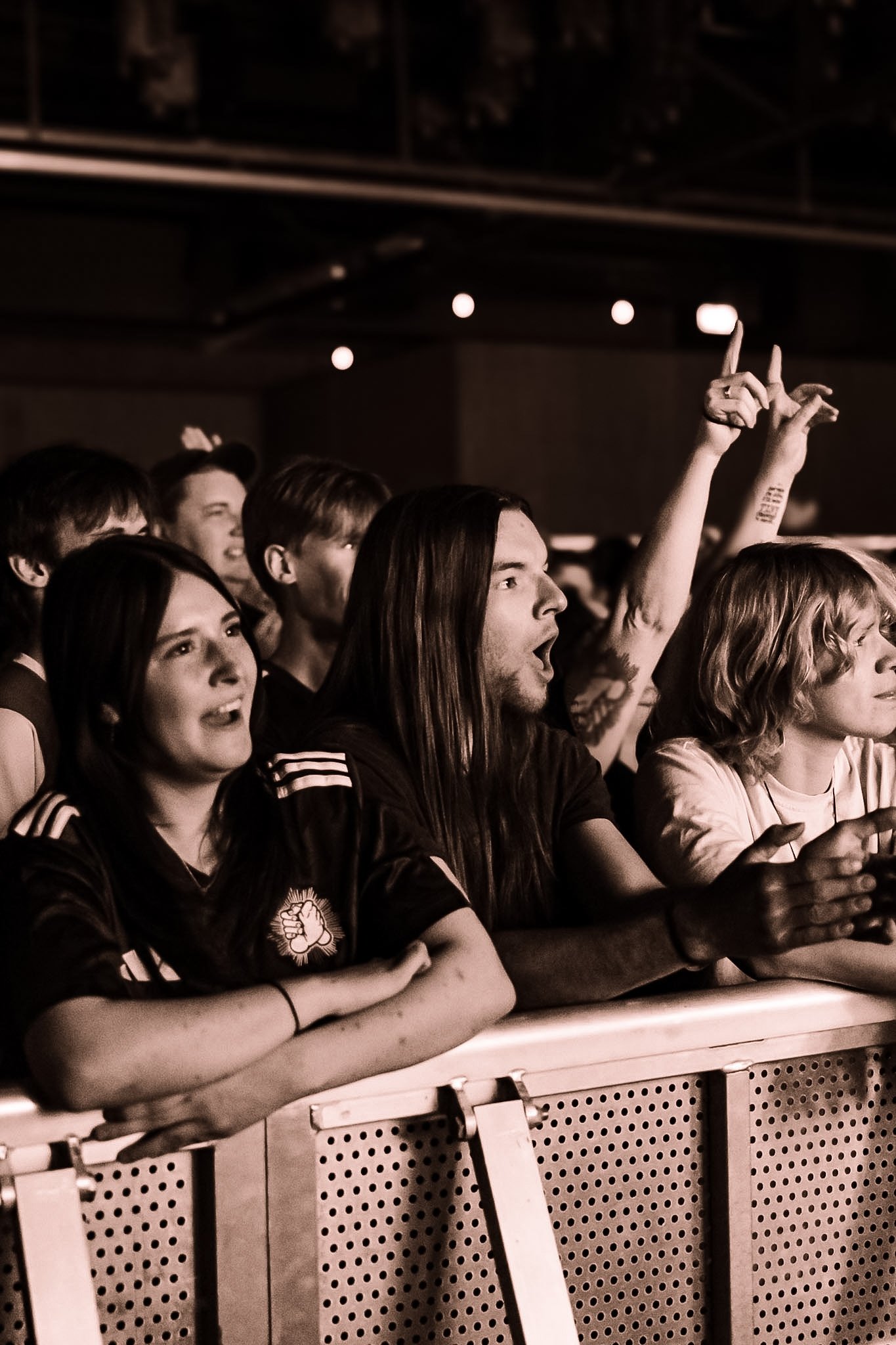 A group of people standing behind a metal barrier at a concert, some with arms raised, exhibiting expressions of excitement and enjoyment.