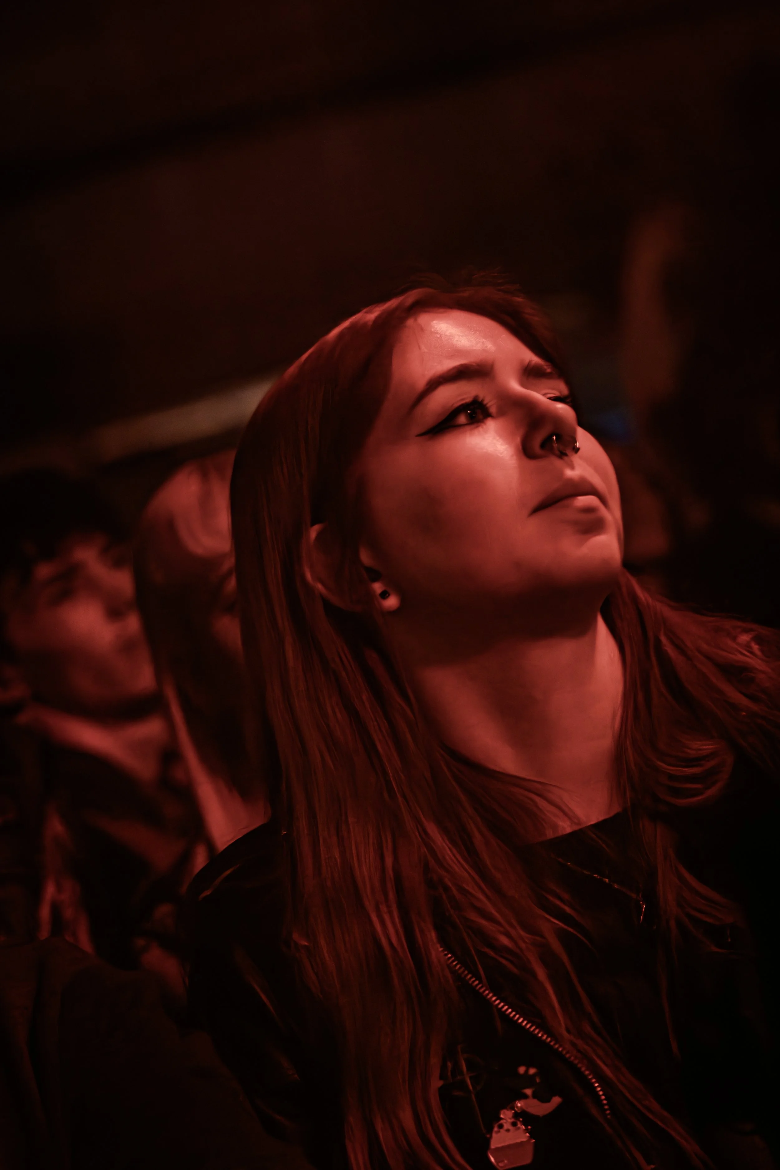 A woman with long red hair, wearing a black jacket, looking upward in a dark setting with red lighting.