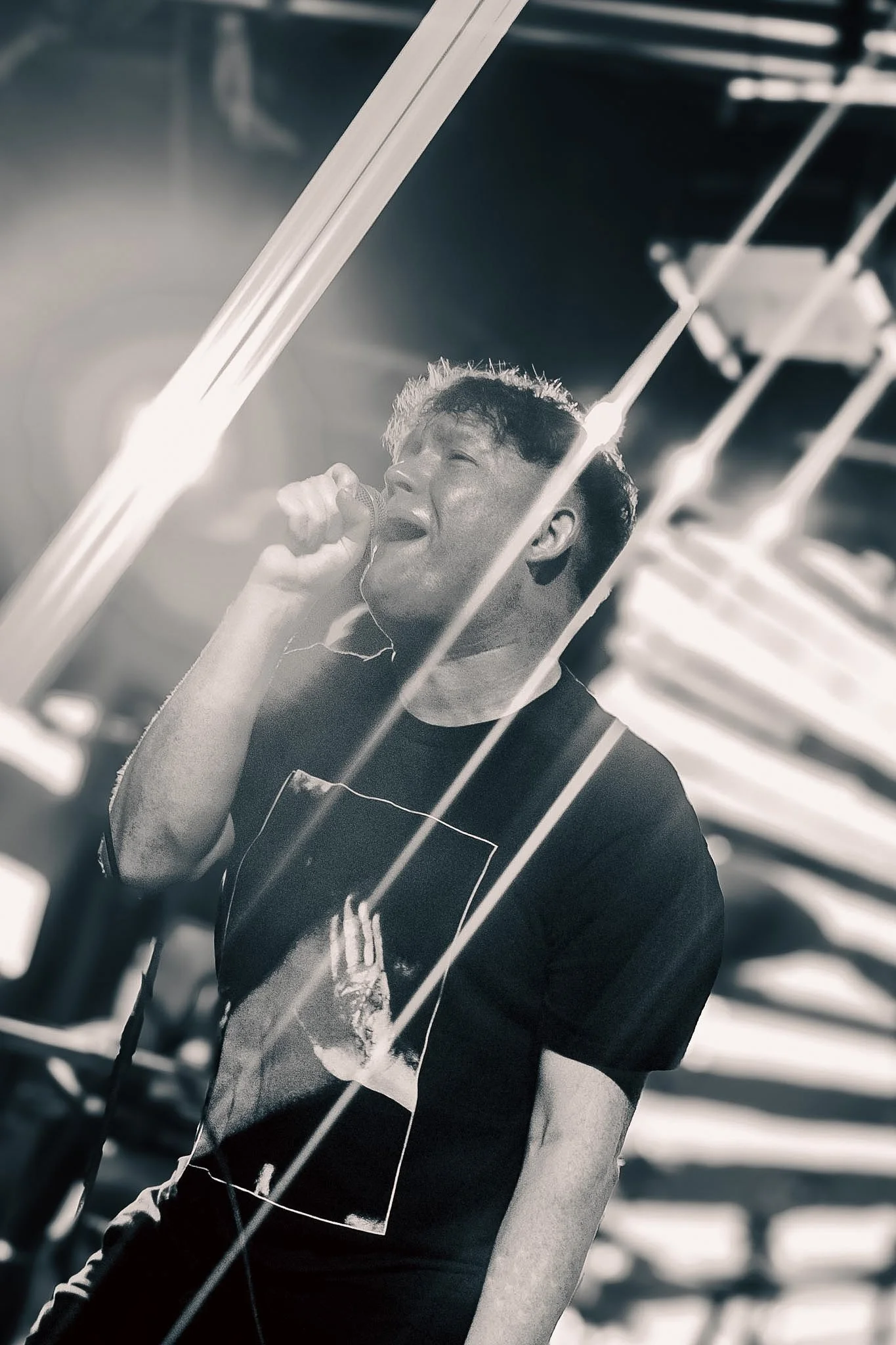 A young man singing into a microphone in a recording studio with musical equipment and bright lights in the background.